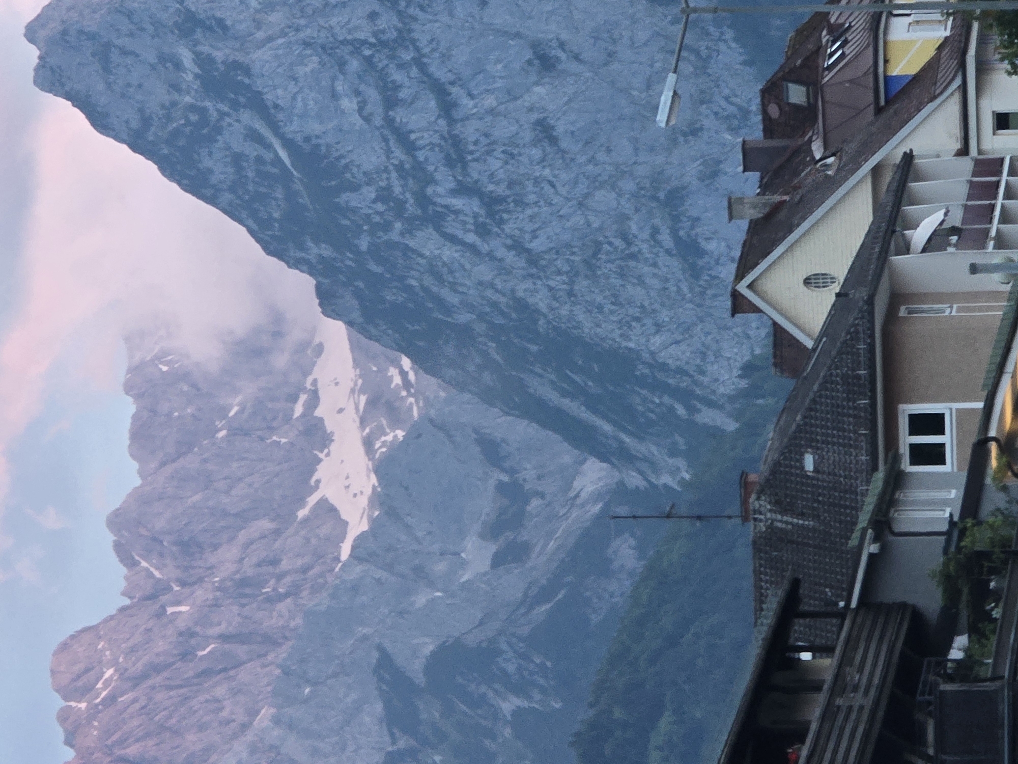 The image shows a view of the Zugspitze massif, Germany's highest peak, from the town of Garmisch-Partenkirchen. In the foreground, several buildings with tiled roofs and balconies are visible. A street lamp is positioned on the right side of the frame. The mountains in the background are steep and rocky, with patches of snow on their upper slopes. The sky is a soft pink and blue, indicating either sunrise or sunset. The overall impression is one of a serene, mountainous landscape.