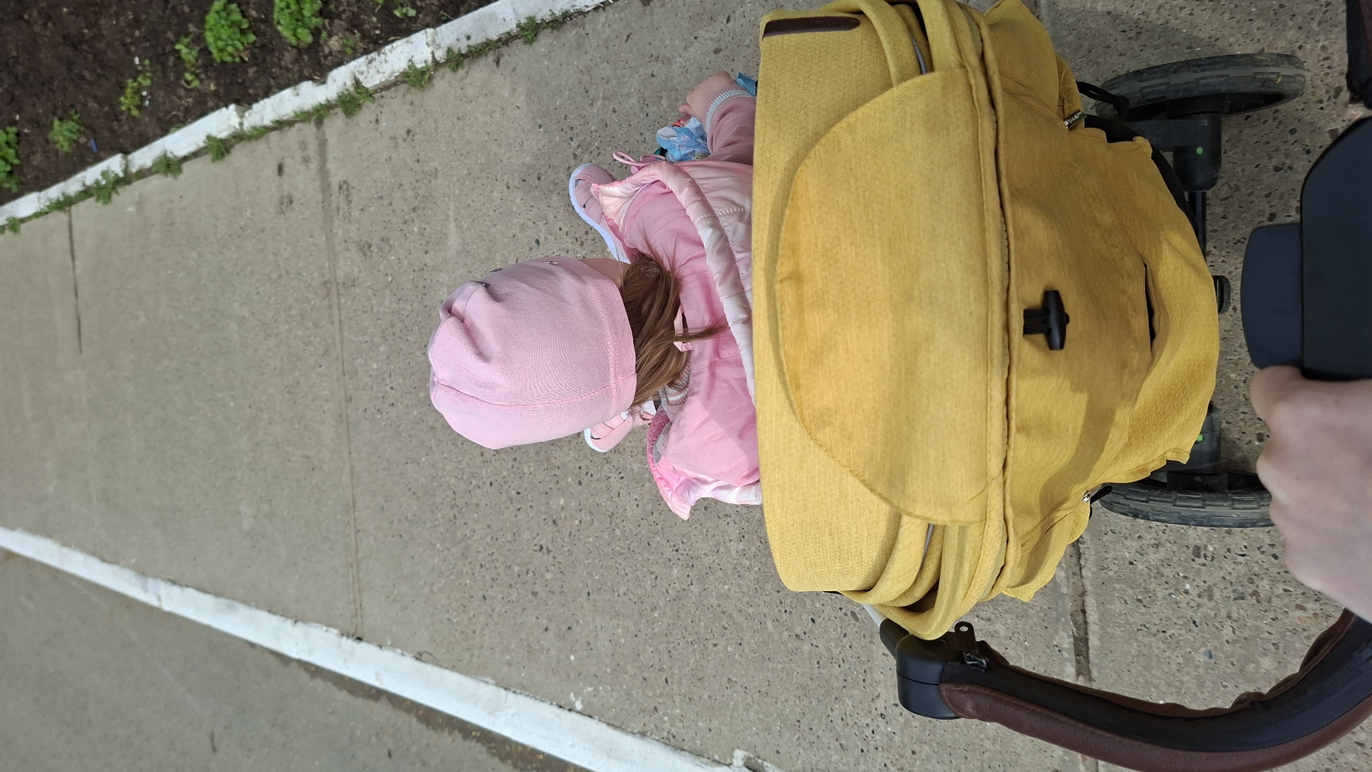 A girl, clad in a pink hat, jacket, and shoes, stands next to a yellow stroller on a concrete sidewalk. The stroller's wheels are visible, and a hand grips the stroller's handle. The setting appears to be an outdoor area in the city of Chaikovsky, Russia.