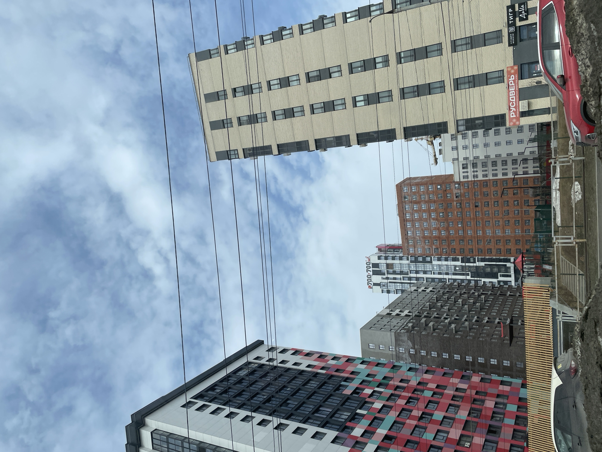 The image shows a street scene in Izhevsk, Russia, dominated by modern high-rise buildings. From bottom to top, the foreground building features a colorful facade with a geometric pattern of red, green, and white squares interspersed with black-framed windows. Behind it are several other residential towers in varying styles: a grey building with protruding balconies, a brown brick building, and a white and grey structure. In the upper part of the frame, there is a beige-colored building with dark-framed windows. A red car is parked on the right side of the photo, next to the beige-colored building. Overhead power lines crisscross the sky, which is partly cloudy. Commercial signage is visible on one of the buildings.
