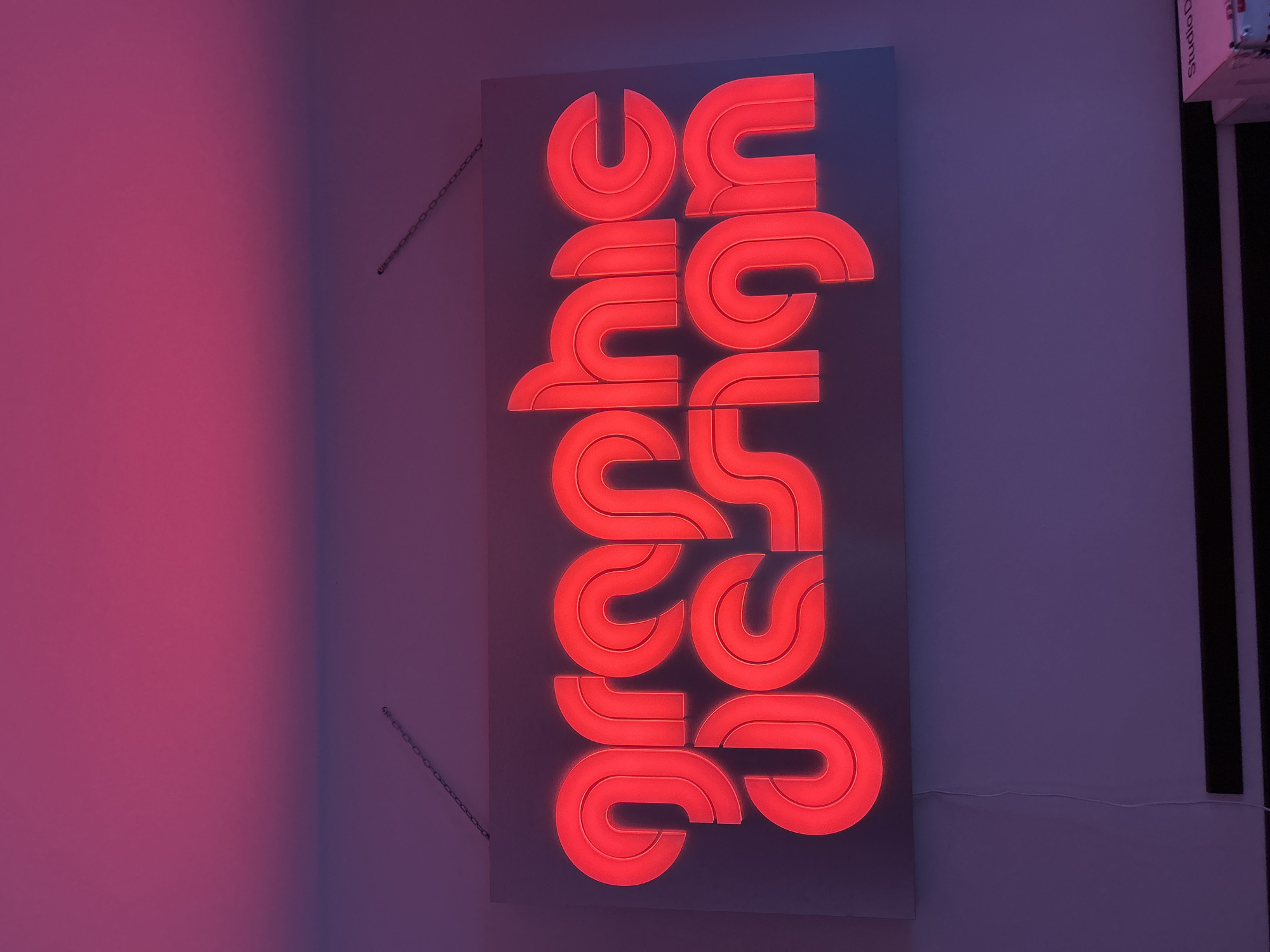 A close-up shot depicts a vertically oriented, illuminated sign that reads "graphic design" in a stylized, geometric font. The letters are formed from a bright red neon-like material that is mounted on a rectangular gray backing. The sign is hanging on a pale purple wall by two small chains. The lighting suggests the presence of a red or purple light source. Part of another sign appears at the upper right corner, "Studio D...". The image was taken in Sofia, Bulgaria.