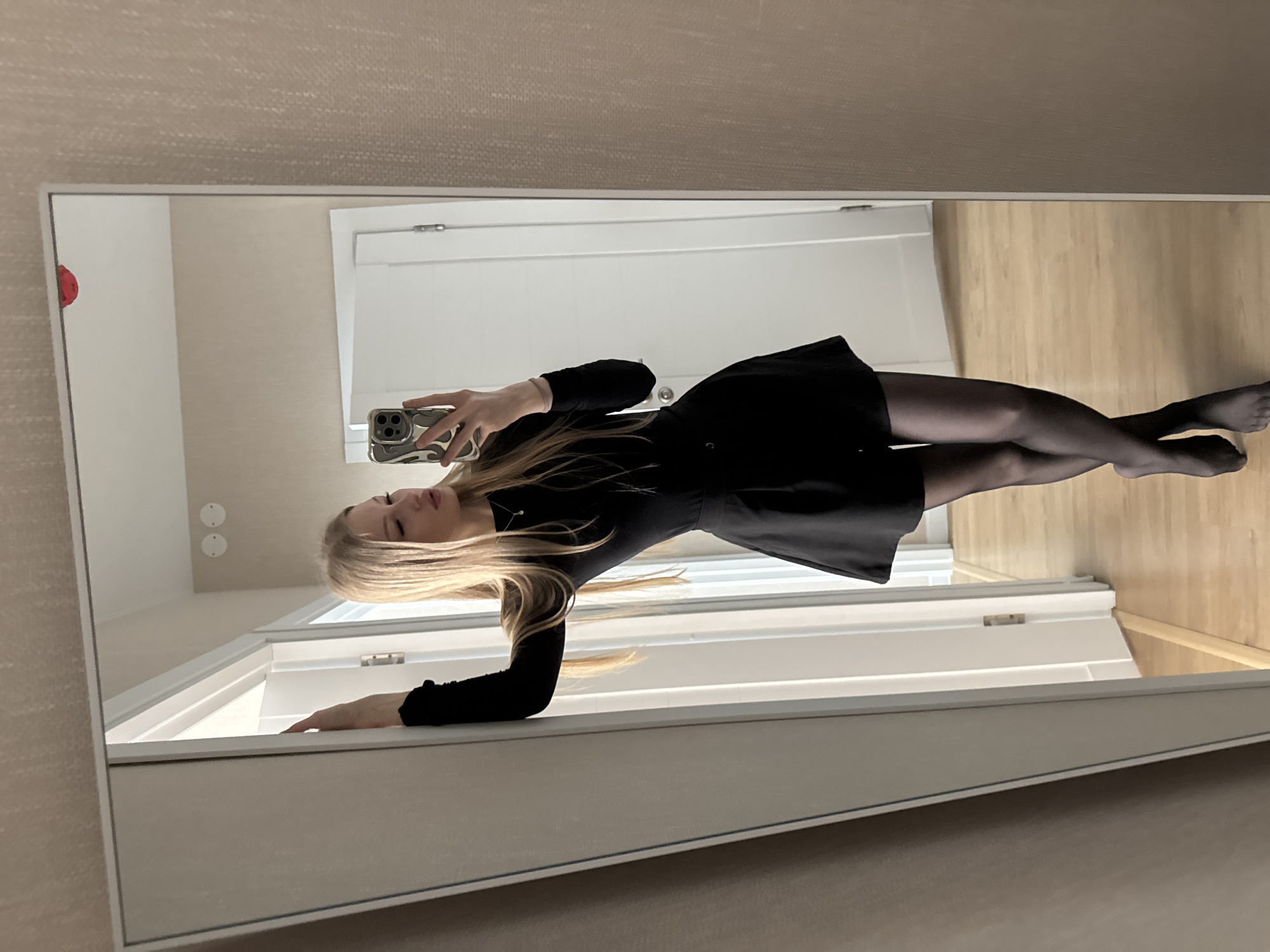 The image displays a full-length mirror reflection of a young adult female in an indoor residential setting. No visual identifiers within the frame allow for the specific identification of the location as Perm, Russia.
The female subject has long, straight blonde hair and light skin. She is dressed in a long-sleeved black top, a black skirt or skort, and sheer black tights, appearing barefoot within the tights. A delicate silver-colored necklace with a small pendant is visible. She stands with her left arm extended upwards, resting on a white vertical surface adjacent to the mirror. Her right hand holds a smartphone, equipped with a distinct black-and-white patterned case, positioned to capture the mirror selfie, partially obscuring her face. Her legs are crossed at the ankles.
The mirror itself is rectangular with a light-colored frame. A small, irregular red object or mark is present on the top edge of the mirror frame. The background reflected in the mirror reveals a light-colored wooden floor, white interior doors with visible hinges, and white walls. Two circular, white ceiling fixtures or elements are also reflected on the ceiling above the doors. The wall immediately to the right of the mirror features light-colored, textured wallpaper. The floor directly in front of the mirror is a matching light-colored wood.