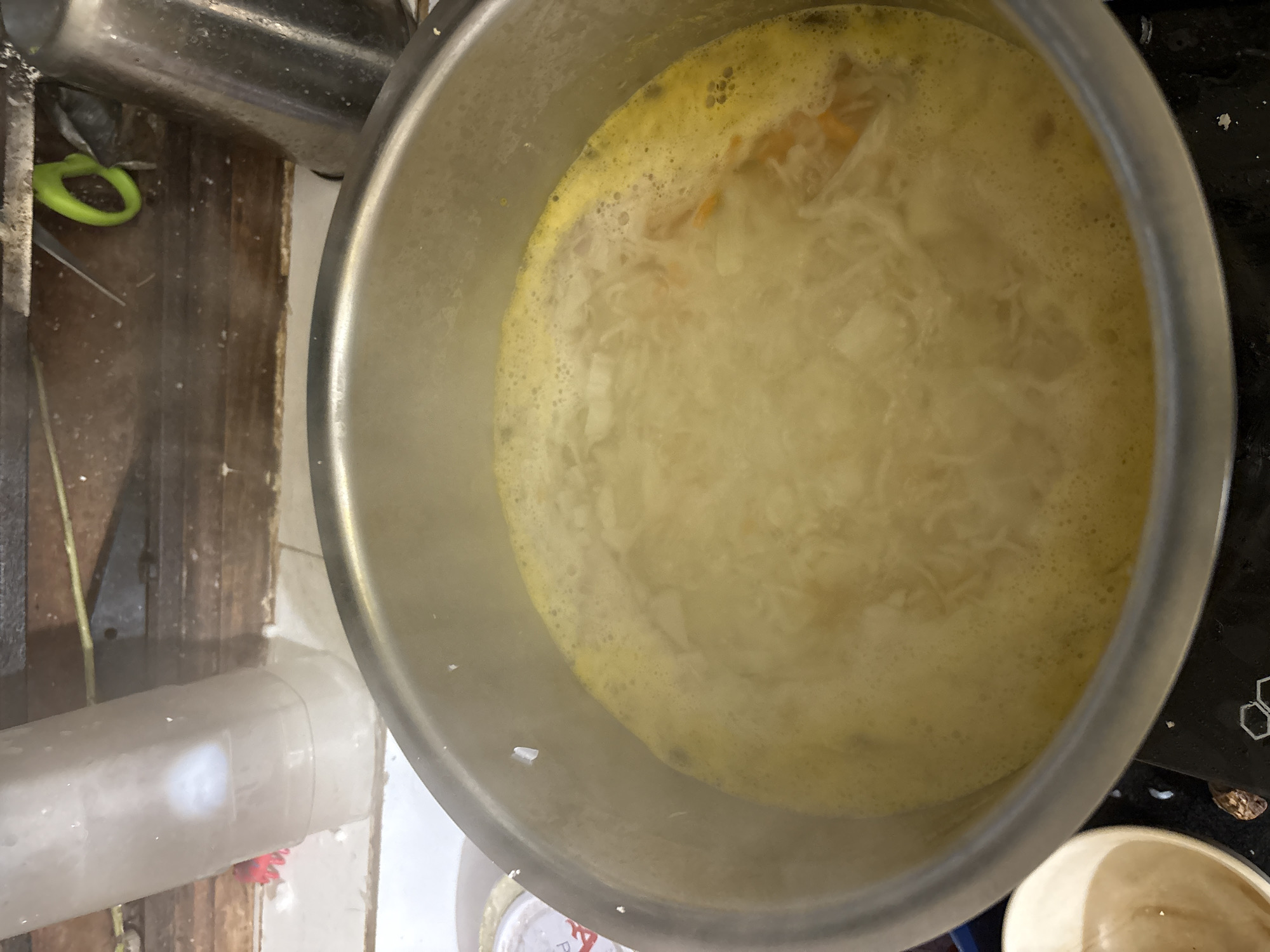 The image shows a close-up, overhead view of a pot of soup simmering on a stovetop. The soup is a pale yellow color and appears to contain shredded vegetables or noodles. The pot is a stainless steel stockpot. In the background, there are wooden shelves with various items, including a clear plastic container, a pair of green scissors, and a metal cup. The setting appears to be a kitchen. There are no people visible in the image. The lighting is somewhat dim, and there is steam rising from the soup. There is no information in the image to suggest the location is Ashvem, India.