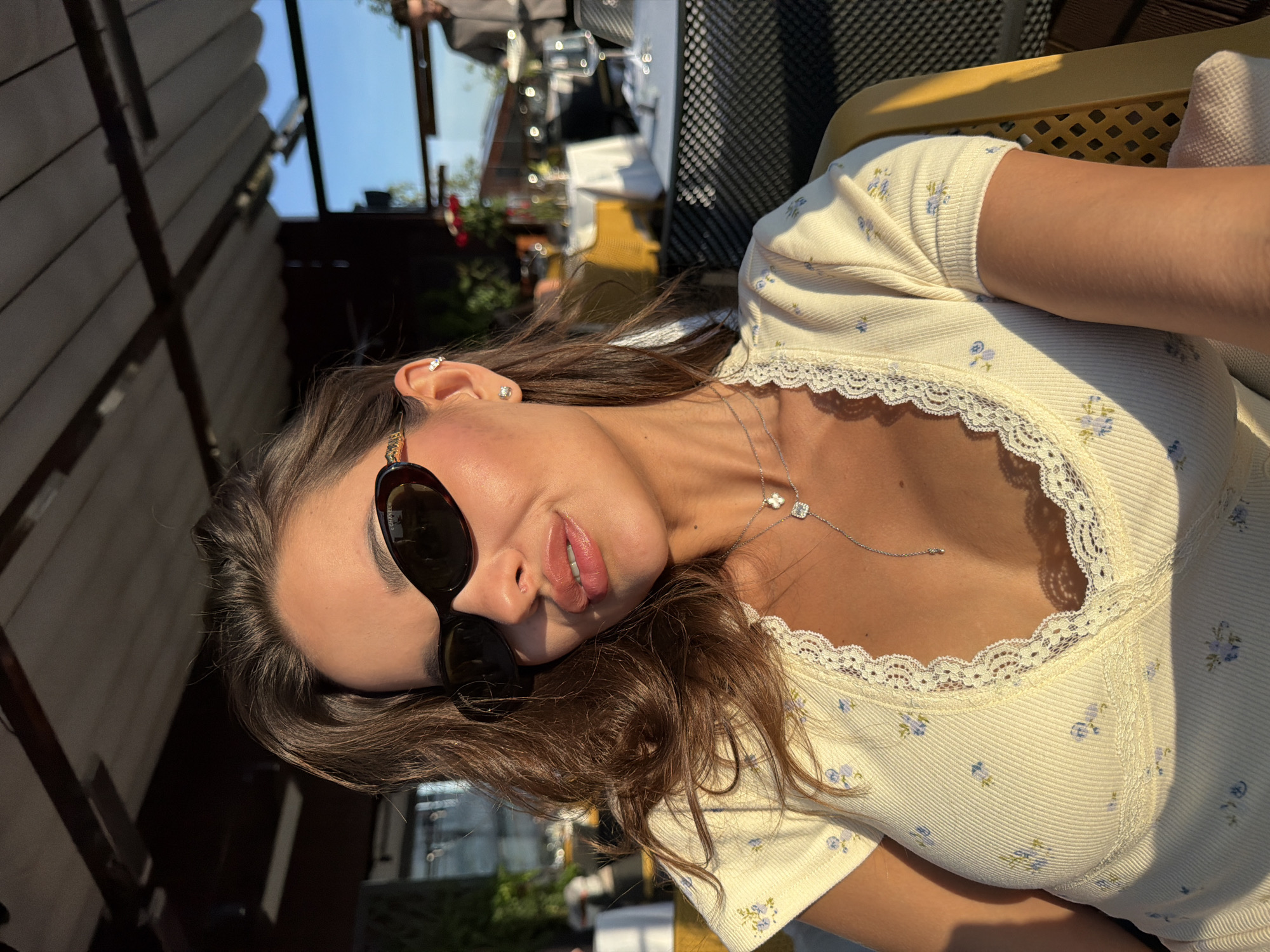 A woman is captured in a close-up, outdoor selfie during sunset. She is wearing a light yellow top with a floral pattern and lace trim, accessorized with sunglasses, a stud earring in her left ear, and a delicate silver necklace. The background suggests a restaurant or cafe setting with tables, chairs, and a covered seating area. The lighting indicates it is late afternoon or early evening. The city of Lednevo, Russia, is not visible in this image.