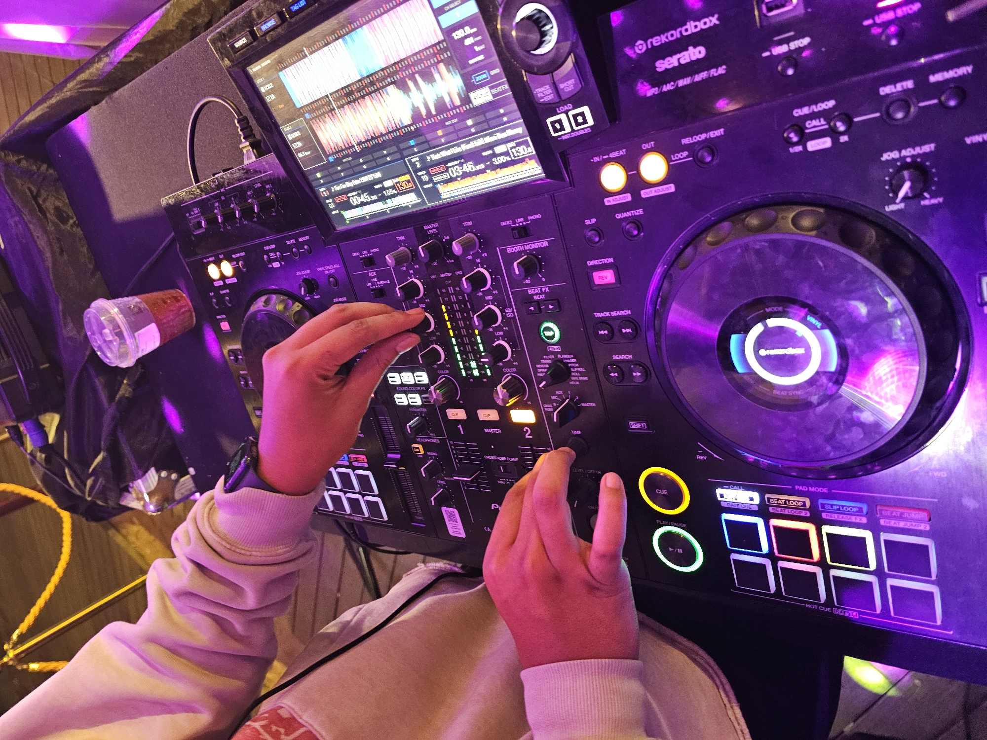 A person, presumably a DJ, is manipulating a black Rekordbox Serato DJ mixing console under purple lighting. Their hands are actively adjusting the mixer's knobs and faders. The console features a central mixer section, jog wheels on either side, and a display screen showing audio waveforms and track information. Various buttons and controls are illuminated, and a disposable plastic cup with liquid is positioned to the left of the console. The environment is dimly lit, and a yellow cable is visible in the periphery. The location, Denpasar, Indonesia, cannot be determined from the visual information.