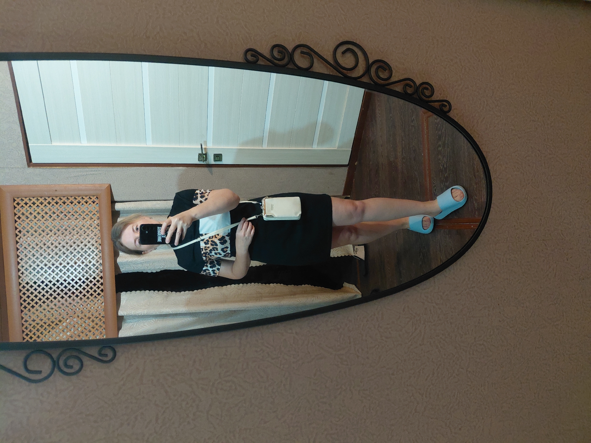 A woman is standing in front of a mirror, taking a selfie. She is wearing a black dress with a leopard print and white detail on the sleeves, a small white crossbody bag, and light blue slippers. The mirror reflects her and part of a room with a door, a curtain, and a decorative screen. The wall has a textured wallpaper. The floor is wooden. The image was taken in a room located in Novosibirsk, Russia.