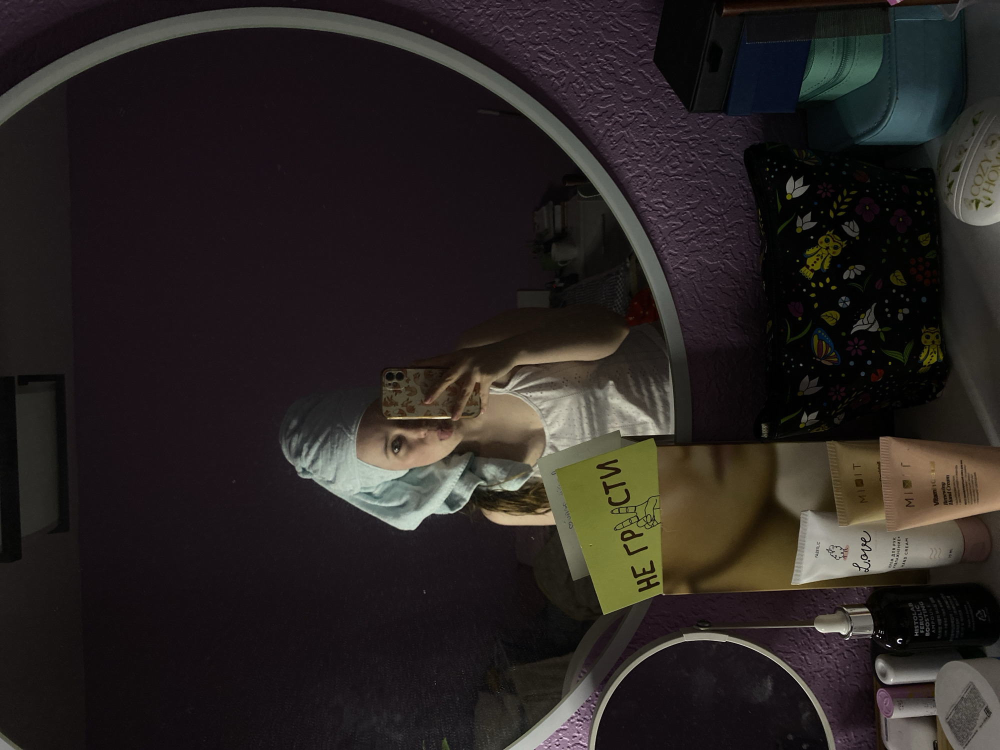 The image displays a mirror selfie taken by a young person, likely female, in what appears to be a personal space such as a bathroom or dressing area. The person is reflected in a round mirror with a light frame, wearing a light-colored, sleeveless top and a light blue towel wrapped around her head, suggesting she has recently washed her hair or is engaged in a post-shower routine. She is holding a smartphone with a floral-patterned case to take the picture, and her lips are slightly puckered, conveying a casual or playful expression.
The background visible in the reflection is a dark purple wall with a textured surface. To the right of the mirror, a shelf or counter holds various personal care products. These include several tubes of hand cream, a dark dropper bottle of an ampoule, and a black cosmetic bag adorned with a pattern of owls and flowers. A white spherical decorative item bearing the text "A COZY HOME" is also present.
A yellow sticky note is prominently displayed on the shelf, facing the mirror. It features the Cyrillic text "НЕ ГРАСТИ" (NE GRASTI), which translates to "DON'T TOUCH" or "DO NOT GRAZE/SCRATCH," along with a drawing of a hand making a peace sign.
Visible text on product labels includes:
- "FABERLIC Love HAND CREAM" (КРЕМ ДЛЯ РУК УВЛАЖНЕНИЕ 70 ML) on a white tube.
- "MIDIT Vitamin C Renewing Hand Cream" on light brown tubes.
- "HISTOLAB FERULIC BOOSTING AMPOULE" on a dark dropper bottle.
The lighting is indoor, suggesting the photo was taken in the evening or morning during a personal grooming ritual. The overall scene captures an intimate and candid moment.