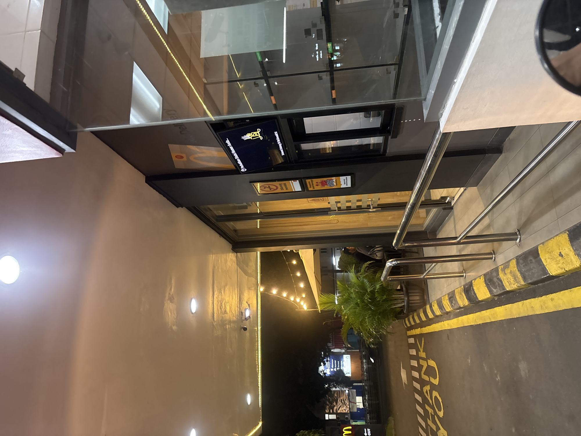 The image depicts the exterior of a McDonald's restaurant at night, seen from a slightly elevated perspective, possibly from inside a vehicle or on an adjacent pathway. The scene is well-lit by artificial lights, suggesting an active business after dark.
In the immediate foreground on the right, a portion of a car's rearview mirror is visible. The ground features a dark asphalt surface with a yellow and black striped curb and yellow painted road markings, including the partial text "CHECK OUT". Metal railings run parallel to the pathway, beside which a lush green potted plant adds a touch of nature. A person is faintly visible standing or sitting near these railings.
The central focus is the modern McDonald's building, characterized by a large glass facade. Reflections on the glass show elements of the interior and surrounding lights. A prominent screen inside displays the golden McDonald's arch logo and text that reads "Perhatikan Disini Nomer Anda...." (Pay Attention to Your Number Here...) and "Selamat Datang Di McDonald's!" (Welcome to McDonald's!). Below this, another sign clearly states "Dilarang merokok" (No Smoking).
Beyond the building and visible through the glass, the dark night sky is illuminated by string lights overhead, creating a festive ambiance. Other distant buildings and lights, along with another McDonald's "M" logo, are visible further in the background. The left side of the frame shows a light-colored wall with several bright, circular light fixtures. The overall setting is consistent with a bustling late-night service point in South Tangerang, Indonesia.