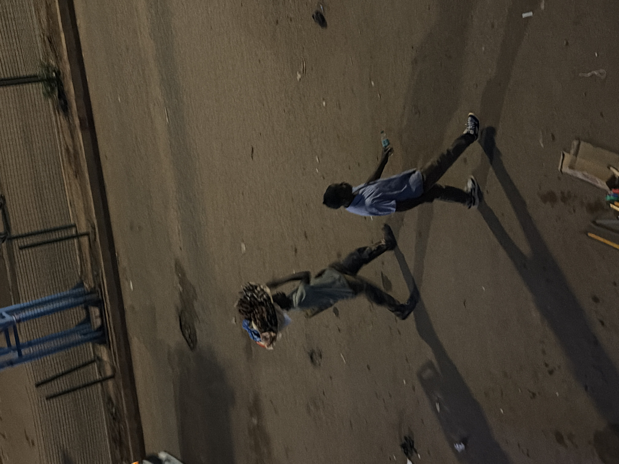 Two individuals are depicted on a paved road, likely in Bukuru, Nigeria, as indicated by the context. The individual on the left, a male, is walking and balancing a load of goods on his head. He is wearing a light-colored t-shirt and dark pants. The individual on the right, also a male, is walking and holding a plastic bottle in his right hand. He is dressed in a light blue short-sleeved shirt and dark pants. The road surface appears worn and somewhat uneven, with scattered debris. In the background, a chain-link fence and some structural elements, possibly part of a building or infrastructure, are visible. The scene is illuminated by artificial light, casting long shadows on the road.