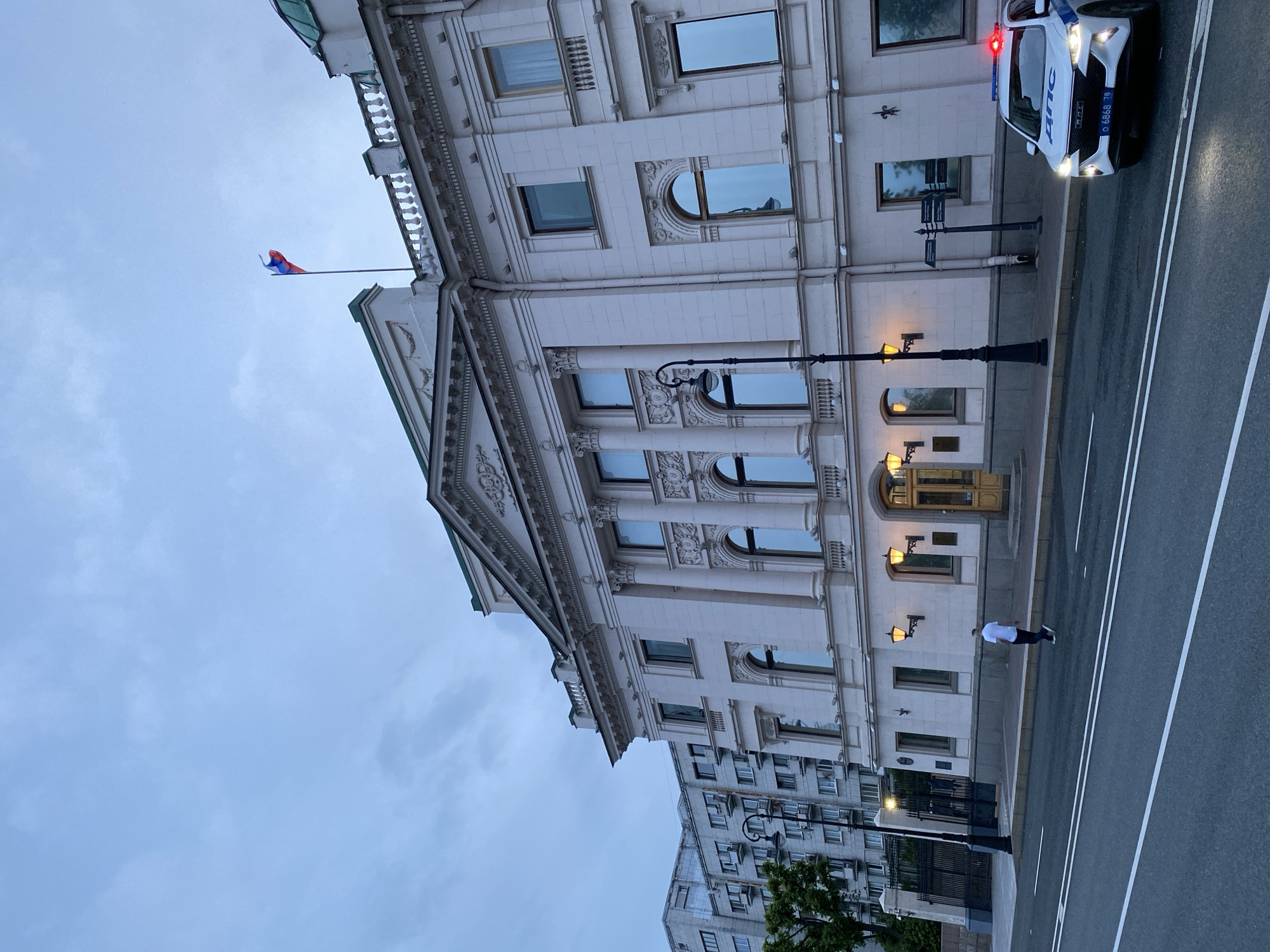 The image shows a street scene in Saint Petersburg, Russia. The foreground is dominated by a wide, paved road with white lane markings. To the left, a man in a white t-shirt and dark pants walks along the sidewalk in front of an ornate, light-colored building. The building features classical architectural elements such as columns, detailed carvings, and multiple windows, some of which are arched. The building's facade is well-lit by several exterior lamps.
Further down the street, a white police car with flashing red and blue lights is parked. The car has Russian license plates and markings. In the background, another building, appearing to be an apartment complex, is visible. The sky is overcast, suggesting a cloudy day, and the lighting indicates it is either early morning or late evening. A Russian flag is visible on a flagpole extending from the main building, adding a national context to the scene. The overall atmosphere is calm, with the presence of the police car being the most dynamic element. The image is oriented vertically, with the street and sidewalk running diagonally across the frame.