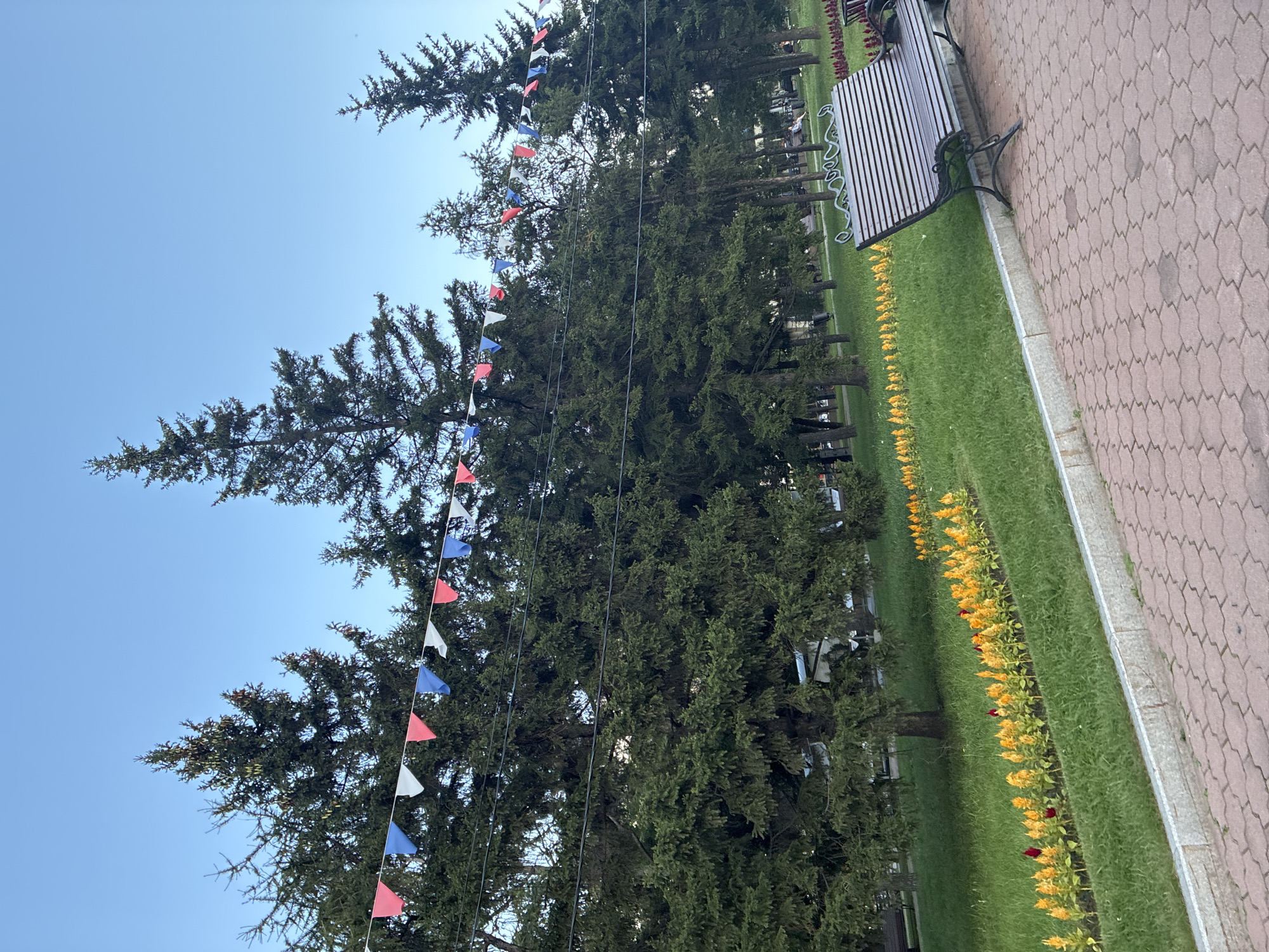 The image captures a park scene in Irkutsk, Russia. In the foreground, a paved walkway with red interlocking pavers leads the eye towards a dark wooden park bench with ornate metal legs. To the left of the bench, a strip of green grass is bordered by a low concrete curb. A row of yellow and orange flowers with red accents is planted along the grass strip. Behind the grass, a dense line of tall evergreen trees stands against a clear blue sky. Decorative triangular flags in red, white, and blue are strung between the trees. In the background, more trees and a glimpse of other park benches and greenery can be seen, suggesting a public park setting. The lighting indicates it is daytime, likely late afternoon or early evening due to the soft light.