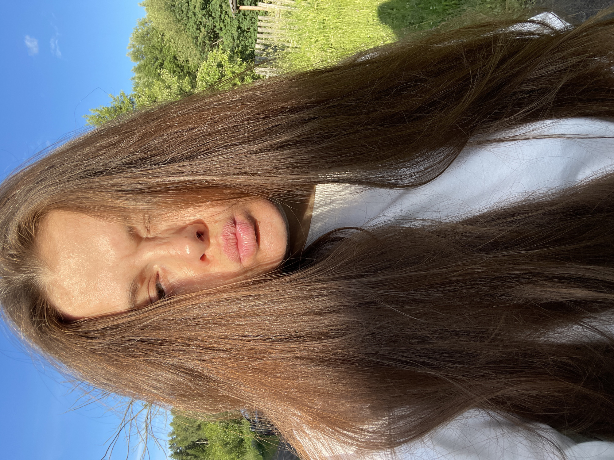 A young woman with long brown hair is taking a selfie outdoors on a sunny day. She is wearing a white shirt and her lips are pursed as if she is blowing a kiss. Her right eye is squinting due to the bright sunlight. The background consists of a clear blue sky, green trees, and a rustic wooden fence. The location appears to be a rural or suburban area, possibly on the outskirts of the city of Zarechny, Russia.