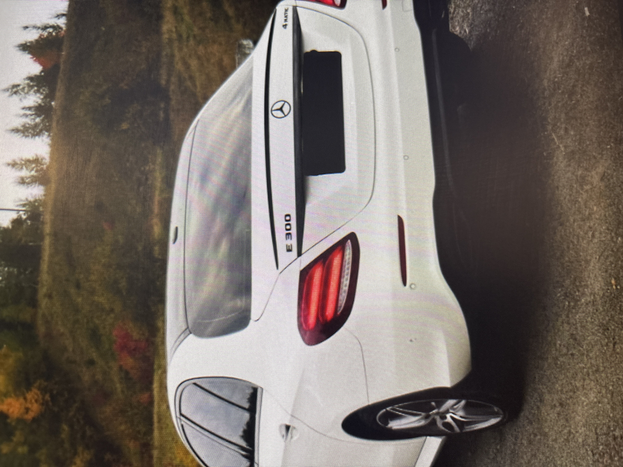 A white Mercedes-Benz E300 4Matic sedan is parked on a gravel road. The car is facing away from the viewer and is angled to the left. The rear of the car is visible, including the taillights, a black license plate cover, and the Mercedes-Benz logo. The car is surrounded by a natural environment with trees and a hillside in the background, suggesting an outdoor location. The image quality is low, with visible pixelation. There is no indication of the location being Moscow, Russia, nor are there any people or specific actions depicted.