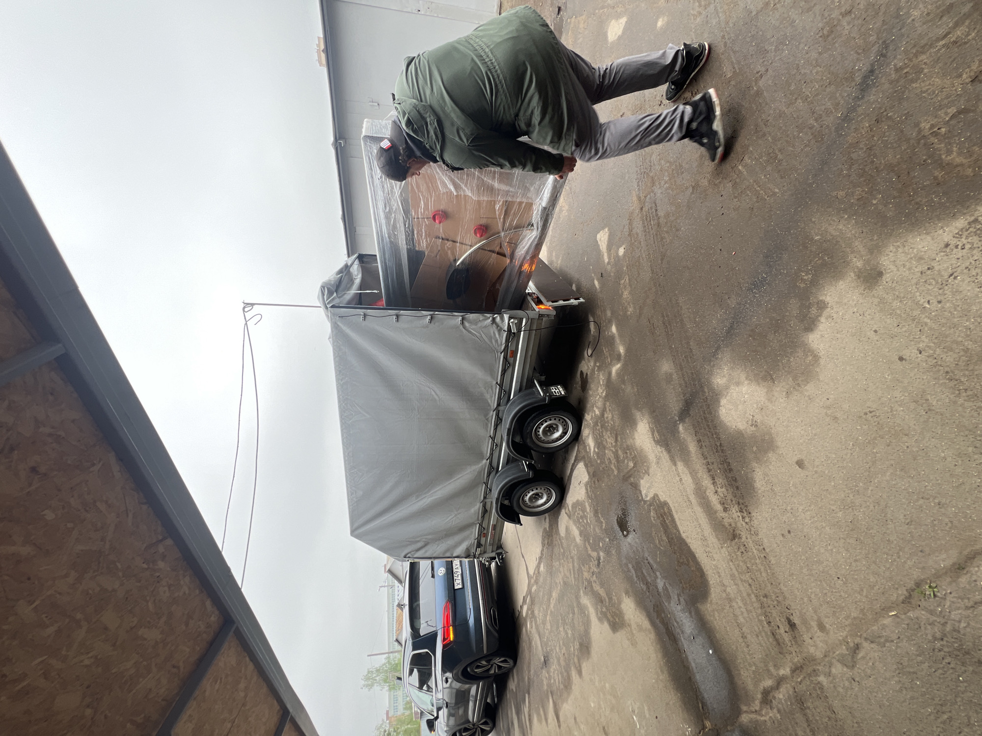 A person is leaning over the back of a trailer, which is attached to a dark-colored car, outdoors. The trailer has a gray cover, and items wrapped in clear plastic are visible at the rear. The person wears a green jacket, gray pants, and black shoes. The background includes buildings, one with light-colored siding and another with plywood siding. The ground is concrete with water marks. A license plate is visible on the car. The location is indicated as Mytishchi, Russia.