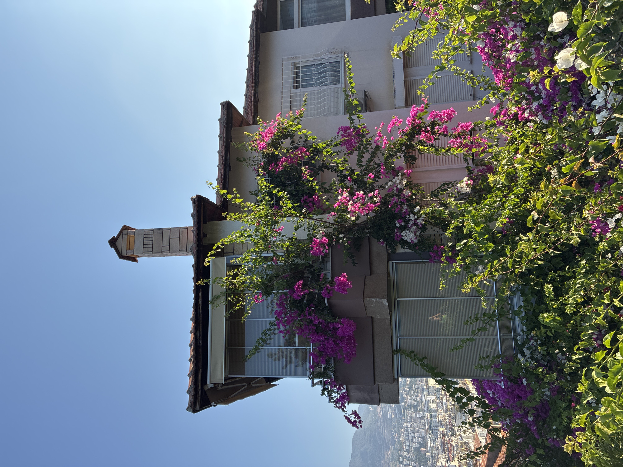 In Alanya, Turkey, this image captures a multi-story building, possibly a residence, adorned with vibrant bougainvillea flowers. The building features a tiled roof and a stone chimney. Two balconies are visible, one on the upper floor with a retractable awning and sliding glass doors, and another on the floor below, also with sliding glass doors. Both balconies are overflowing with pink and white bougainvillea. The lower portion of the building is obscured by a dense arrangement of green foliage and more bougainvillea. In the background, a cityscape with numerous buildings stretches across a hillside, indicating an urban environment. The sky is clear and blue, suggesting a sunny day.