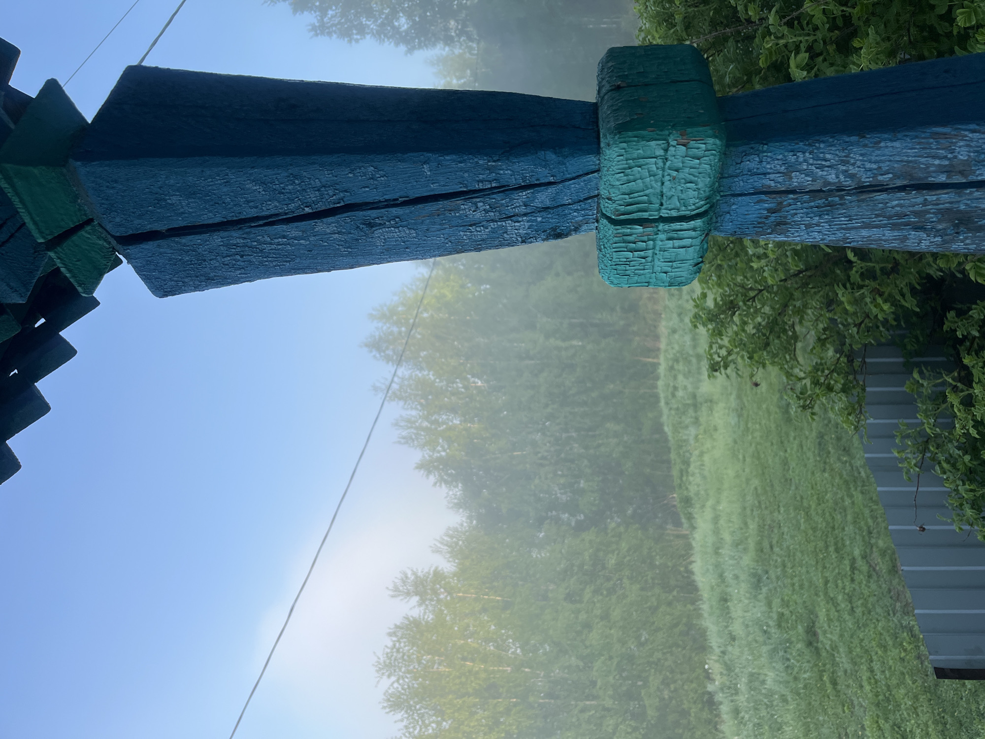 The image shows a section of a painted wooden structure, possibly a gazebo or porch roof, with blue-green paint, set against a background of trees, grass, and a fence. The structure's paint is peeling or weathered. A light fog obscures the trees in the background. The location is possibly in a rural or suburban area, and given the request, potentially Nizhny Tagil, Russia. There are no people visible in the image.