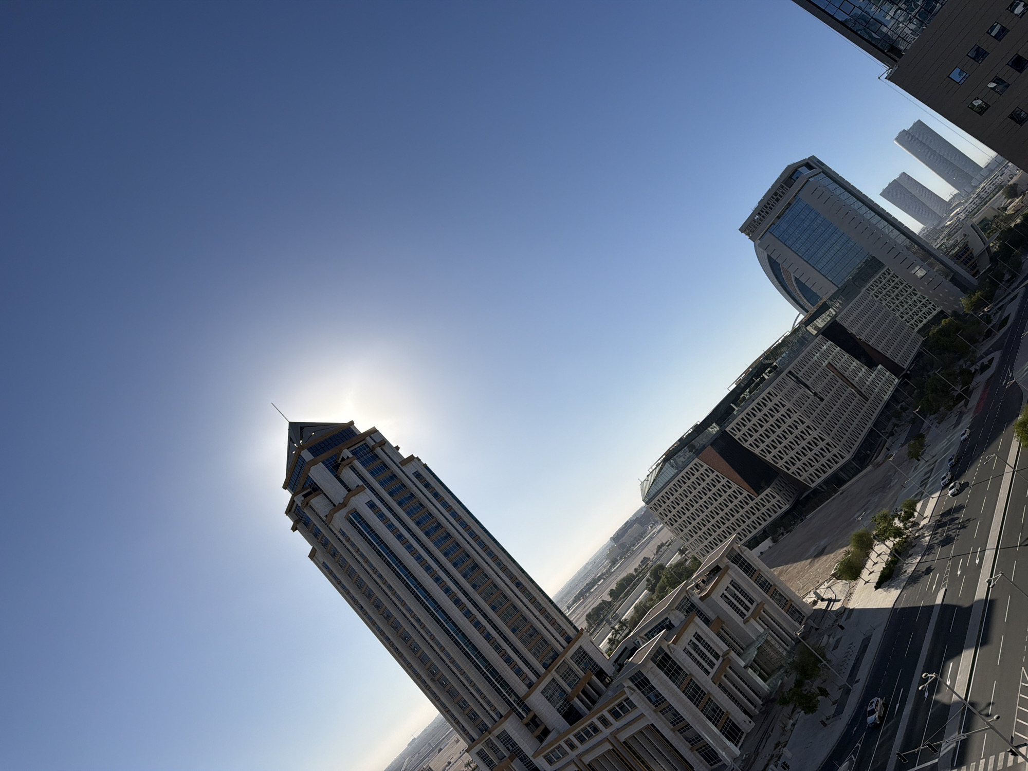 The image shows a cityscape dominated by modern architecture under a clear blue sky. In the foreground, a tall, slender skyscraper with a distinctive pointed roof and light-colored facade is angled upwards, with the sun partially obscured behind its peak. The skyscraper has numerous windows arranged in vertical and horizontal lines.
To the right of the main skyscraper, two distinct buildings are visible. One is a low-rise structure with a glass facade and a patterned exterior. Adjacent to it is a taller, curved building with a glass front. Further in the distance, several more skyscrapers rise, their forms softened by atmospheric perspective.
The lower portion of the image reveals a street scene with cars driving on a multi-lane road. Trees line the sidewalk, and shadows are cast by the buildings, indicating a bright, sunny day. The overall impression is one of urban development and modern infrastructure. The image appears to be taken from an elevated vantage point, looking down on the street and across the city.