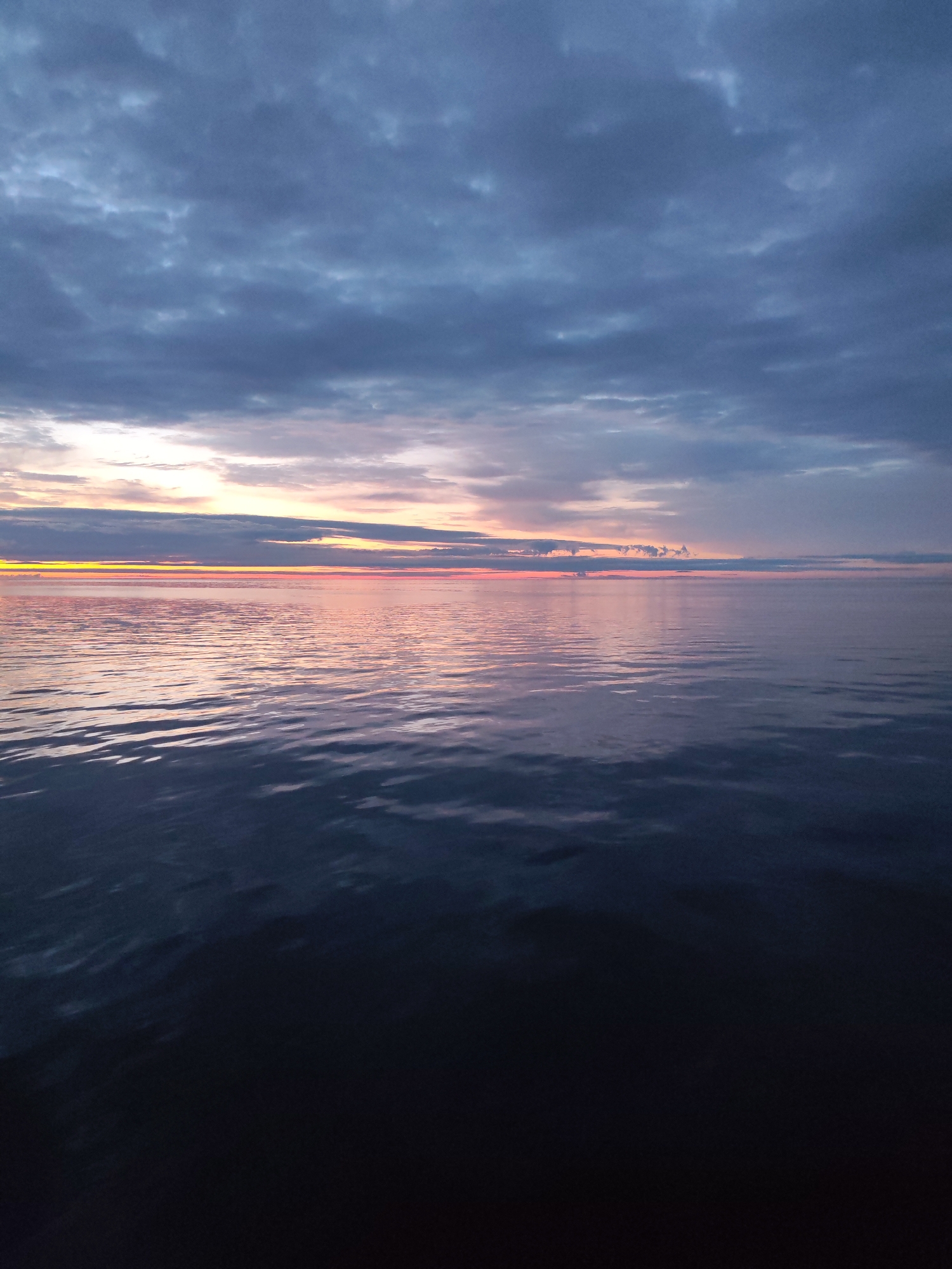 The image depicts a serene seascape at dusk, likely near Shcheleiki, Russia. The sky is dominated by a palette of deep blues and grays, with scattered clouds tinged with soft hues of pink and orange from the setting sun. A thin band of vibrant yellow and orange light streaks across the horizon, marking the last rays of daylight. The water is calm, reflecting the muted colors of the sky, with subtle ripples creating a textured surface. The foreground of the water appears darker, transitioning into a deep, almost black hue, suggesting the encroaching night. There are no people, structures, or distinct landforms visible, creating a sense of vastness and tranquility.