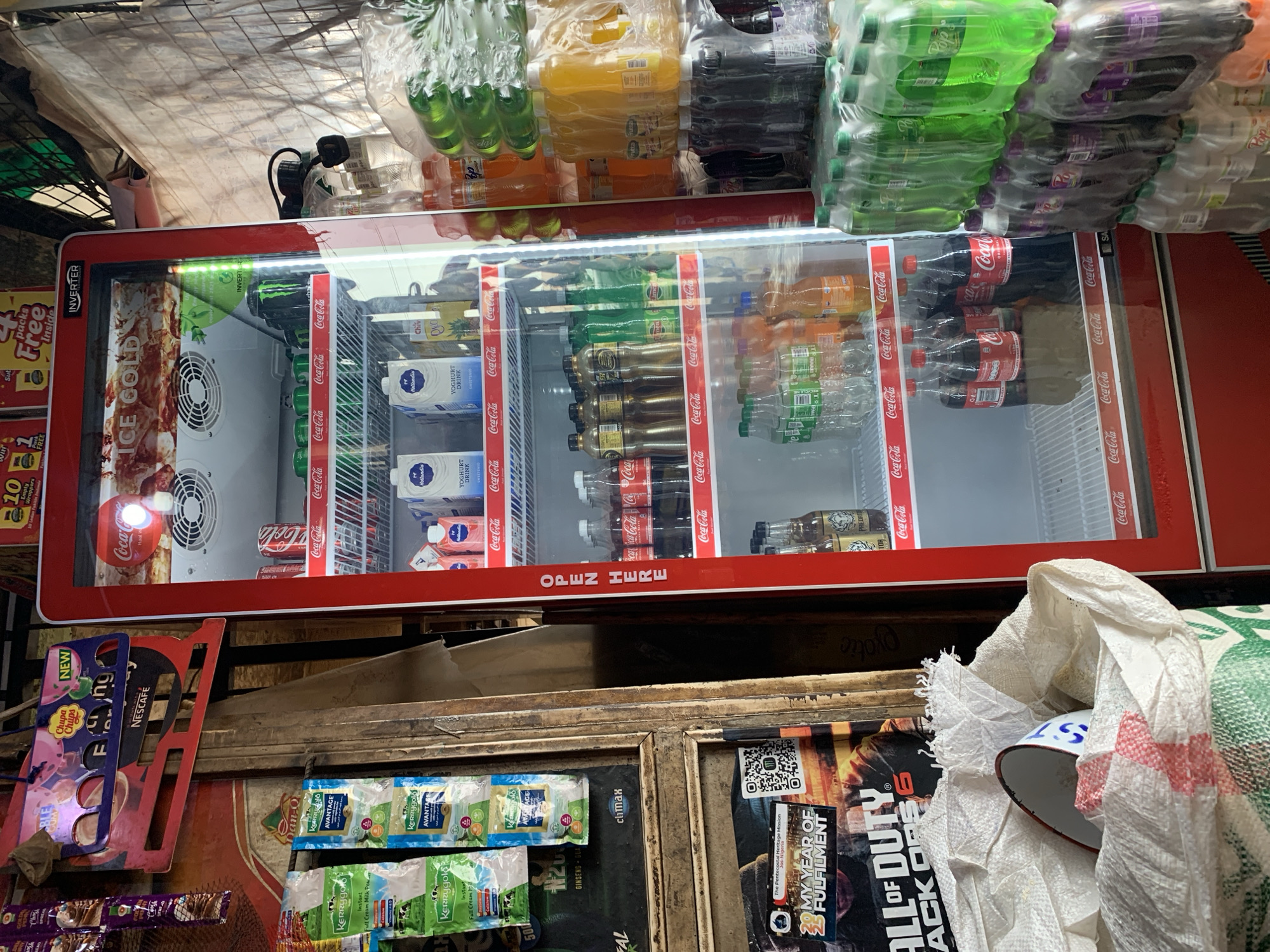 The image shows a refrigerated display case stocked with various beverages, including soda, juice, and yogurt drinks. The case is red and bears the Coca-Cola logo along the edges. The products are neatly arranged on shelves within the case. Above the refrigerator, various packages of drinks and snacks are visible. Below the display case, there are several items, including packages of powdered milk, candy, and a Call of Duty Black Ops 6 poster. The environment appears to be a small shop or kiosk in the city of Rafin Par, Nigeria, possibly a roadside vending spot.