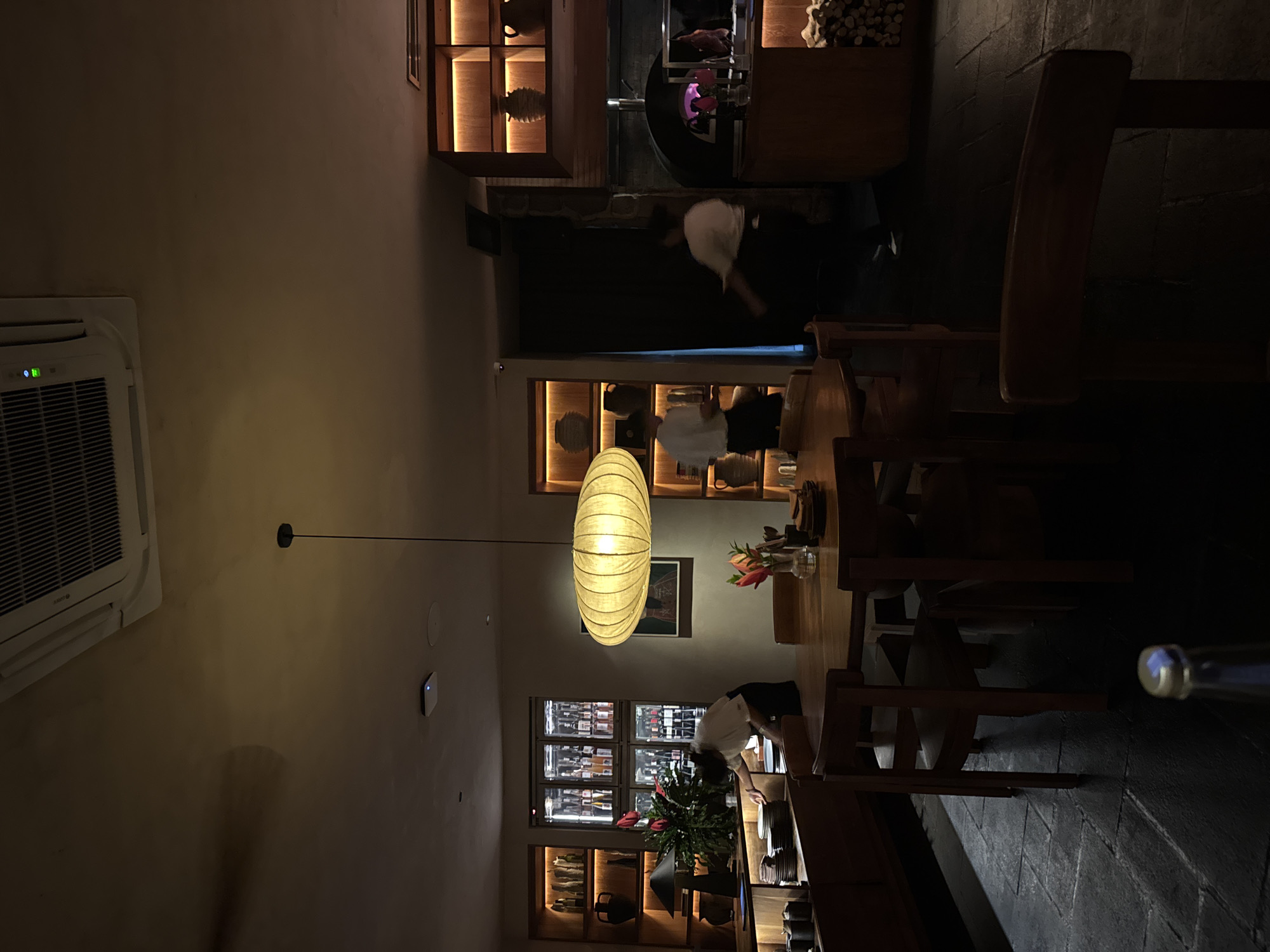 The image captures the interior of a restaurant in North Kuta, Indonesia. The dining area features wooden tables and chairs, illuminated by a large, decorative pendant light. Shelving units, some backlit, display decorative objects and bottles, possibly wine. Waitstaff is present, interacting with the dining space. Visible near the top of the image is a pizza oven. An air conditioning unit is mounted on the wall on the left.