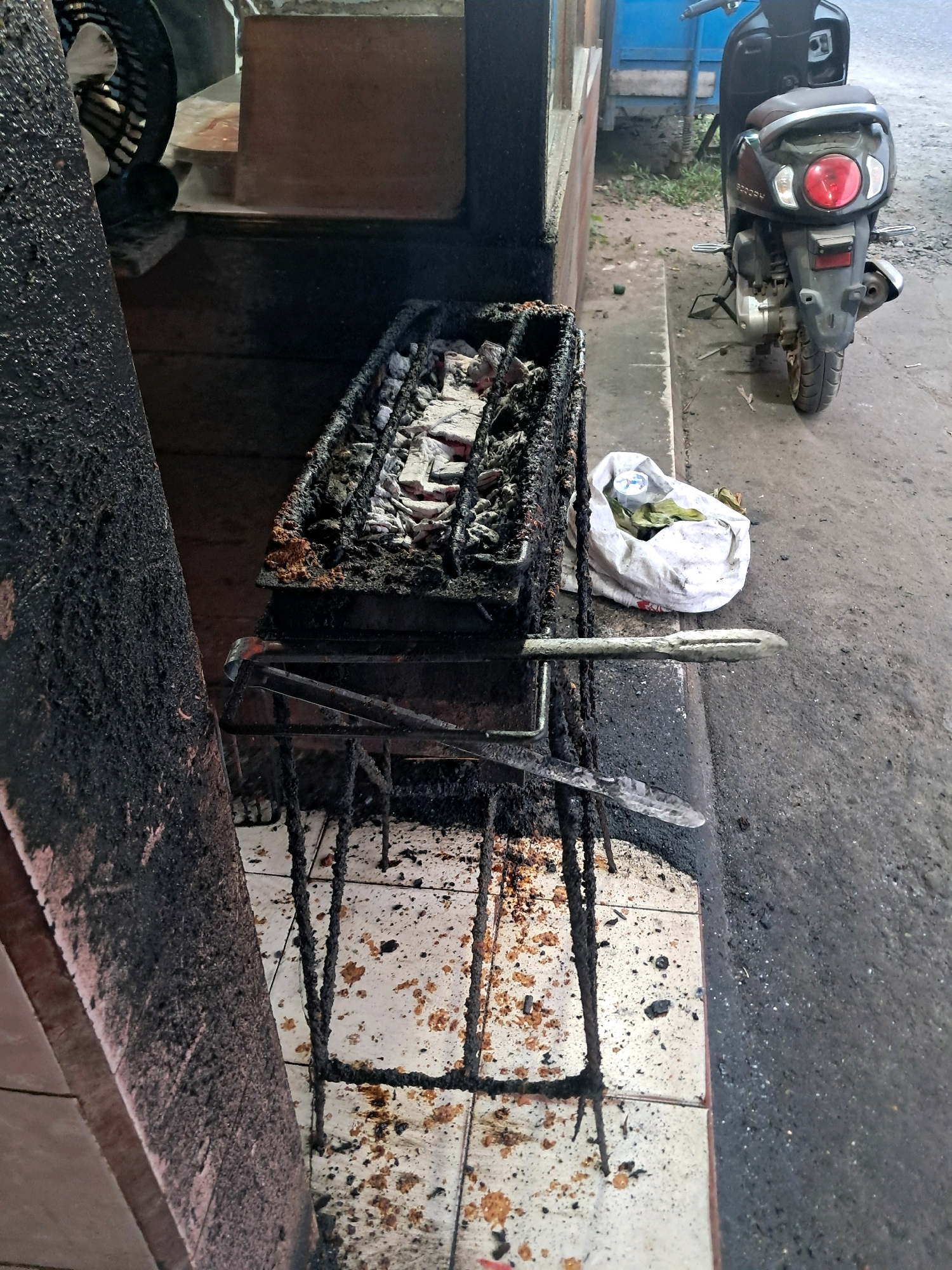 The image shows a charcoal grill with cooking remnants and a white bag of items next to it, placed on a tiled surface. A motorcycle is parked behind the grill. The scene is outdoors, likely in a public area, with a worn paved ground and some vegetation in the background. The grill and surrounding area appear dirty, with grease and ash spread across surfaces. The overall impression is that of a food stall or a food preparation area that is in use or has recently been used.