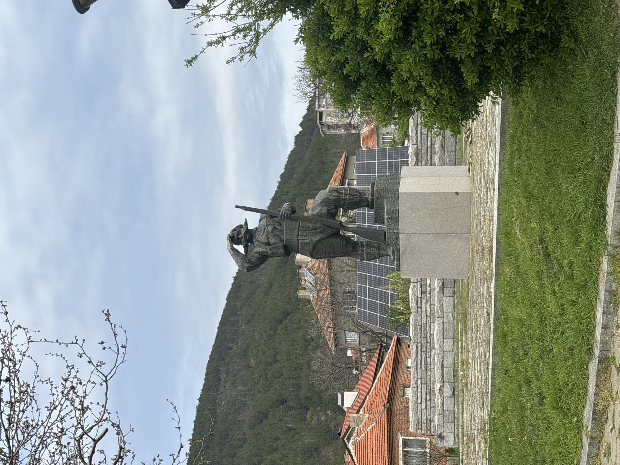 The image captures a bronze statue of a man in military garb, appearing to be gazing into the distance with a hand shielding his eyes. He is holding a long object, possibly a rifle. The statue stands on a stone base, set within what appears to be a small park or public area. The scene includes a backdrop of red-tiled rooftops, suggesting a residential area, and a lush, forested hillside. Additionally, solar panels are visible, indicating a blend of old and new technologies in the environment. The statue and the surrounding landscape suggest an Eastern European setting. The exact location in the city of Unknown, Unknown is unknown.