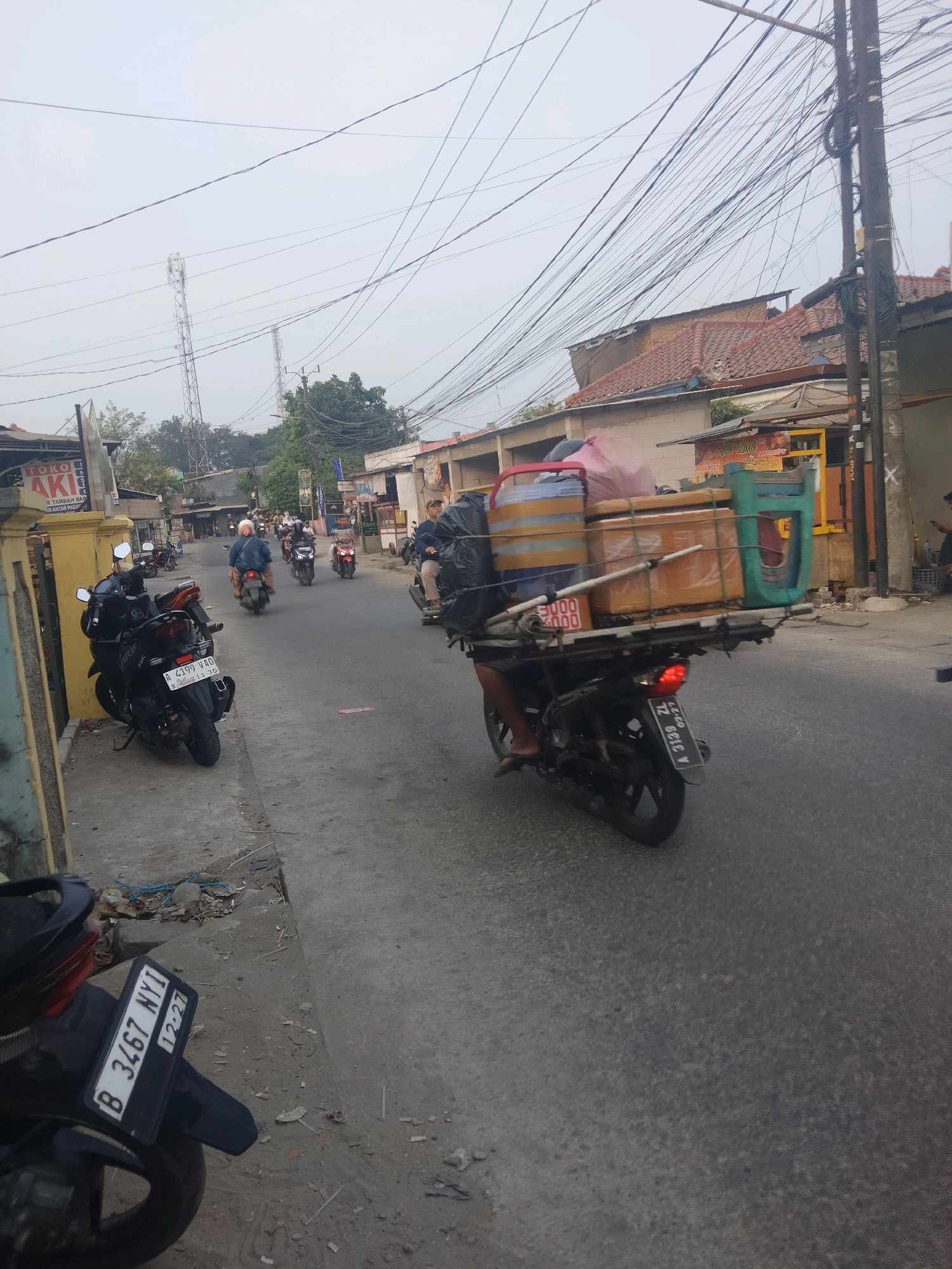 This is a medium shot, eye-level, vertical photograph taken on a street in Suka Sari, Indonesia. The scene depicts a street with moderate traffic, mostly motorcycles. In the foreground, on the left, is a parked black motorcycle with a license plate reading "B 3467 NYI 12-27". Further down the street, a motorcycle with a large load secured to the back is in motion. The load consists of various items, including plastic containers, boxes, and pink bags, all strapped down to a flatbed attached to the rear of the motorcycle. The rider of this motorcycle is wearing a blue shirt and dark shorts. Other motorcycles are visible in the background, some carrying passengers. The street is lined with buildings and houses, some with red tile roofs. Utility poles with numerous wires crisscross overhead, suggesting a somewhat dense urban or suburban environment. The sky is overcast. The overall impression is of a busy street scene with everyday activities and transportation.