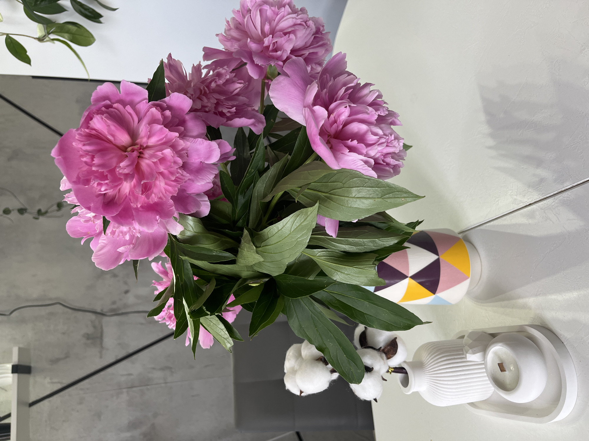 A bouquet of pink peonies is arranged in a colorful geometric vase on a white table. To the left of the vase, on a white tray, are a white ribbed vase, a small white geometric sculpture, and a white candle in a round holder. Behind the bouquet, a cotton branch with fluffy white bolls is visible. The background consists of a concrete wall and a white wall with a plant. The scene appears to be indoors.