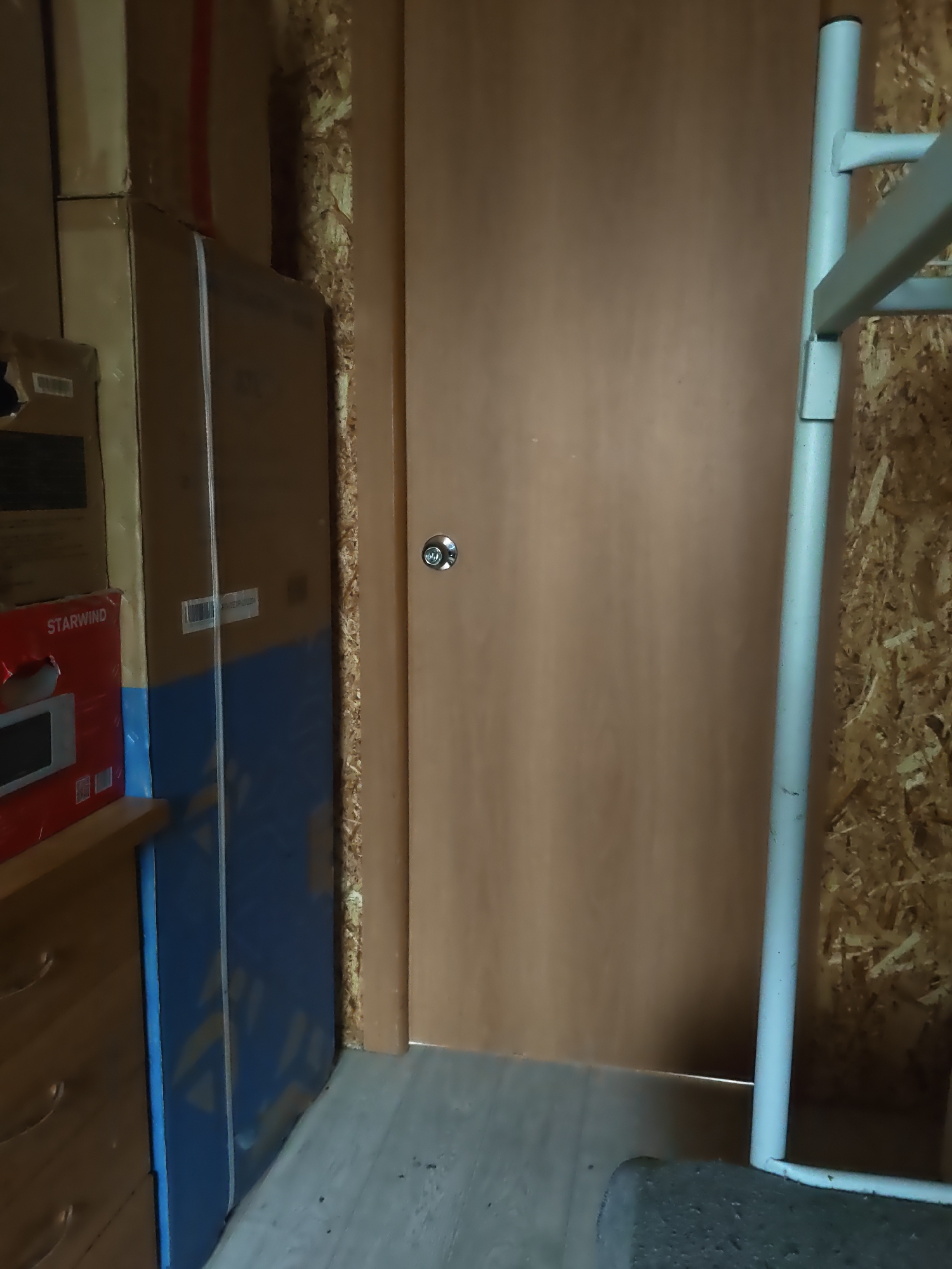 In a dimly lit room, possibly a storage area or shed, a light brown wooden door with a silver doorknob is centrally positioned. To the left of the door, stacks of cardboard boxes are visible. One box, red with "STARWIND" branding, is partially obscured, showing a glimpse of a microwave oven. Next to this, a tall, blue object with a diagonal pattern is present. The floor appears to be made of light-colored laminate. To the right of the door, a light blue metal structure, possibly part of a rack or scaffolding, is present, extending upwards and to the right. The wall behind the metal structure is rough and appears to be made of oriented strand board (OSB). The overall environment suggests an indoor space, likely a residential or commercial storage area, within the city of Platoschino, Russia. There are no people or visible animals in the image.