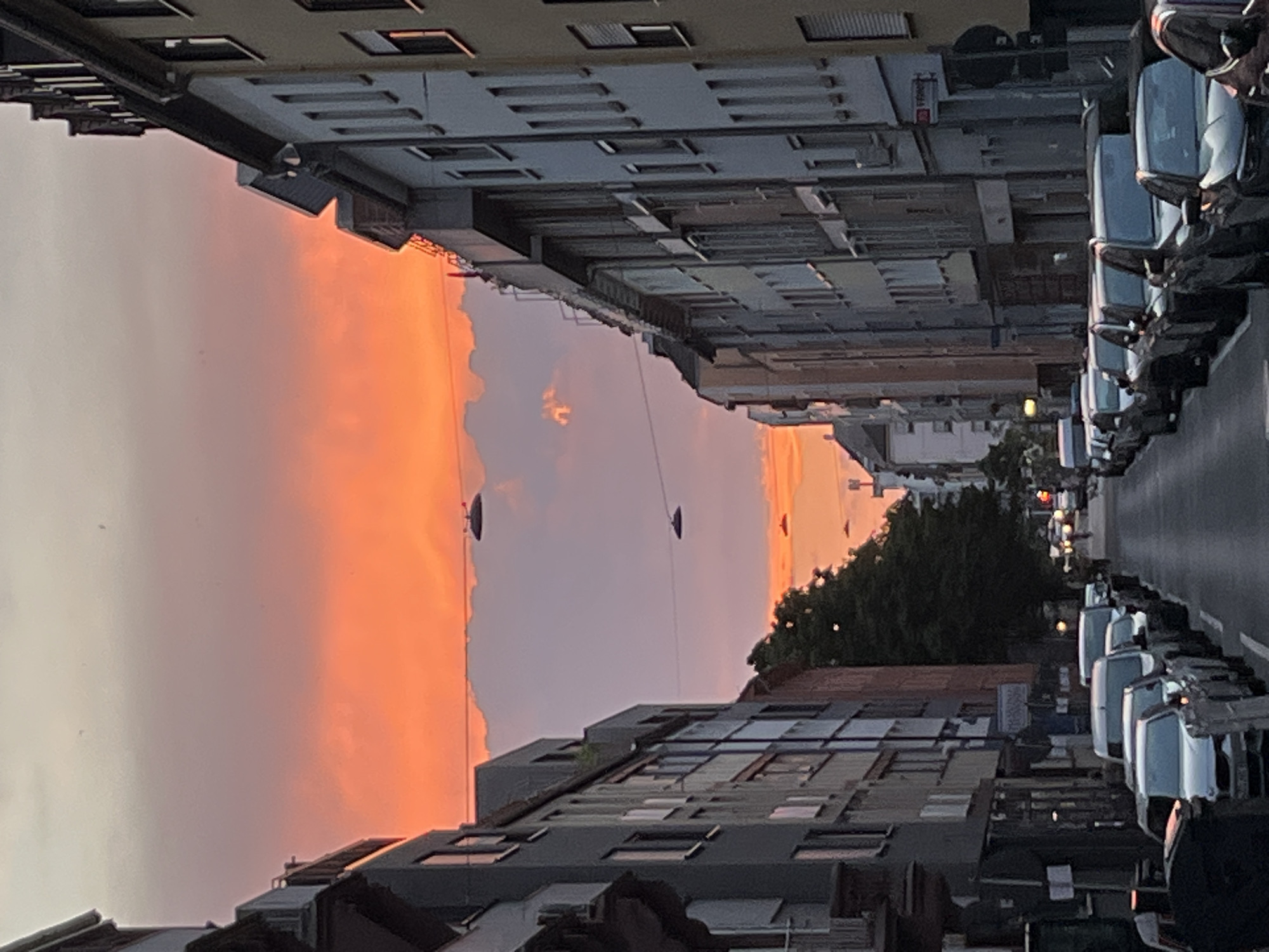The image shows a street scene at sunset in an urban environment. The sky is filled with vibrant orange and pink clouds. Several dark, disc-shaped objects are suspended from thin wires, appearing to be some form of streetlights or decorative lighting. The street is lined with multi-story buildings on both sides, with windows and balconies visible. A row of parked cars is visible along the right side of the street. The lighting suggests it is either dusk or dawn.