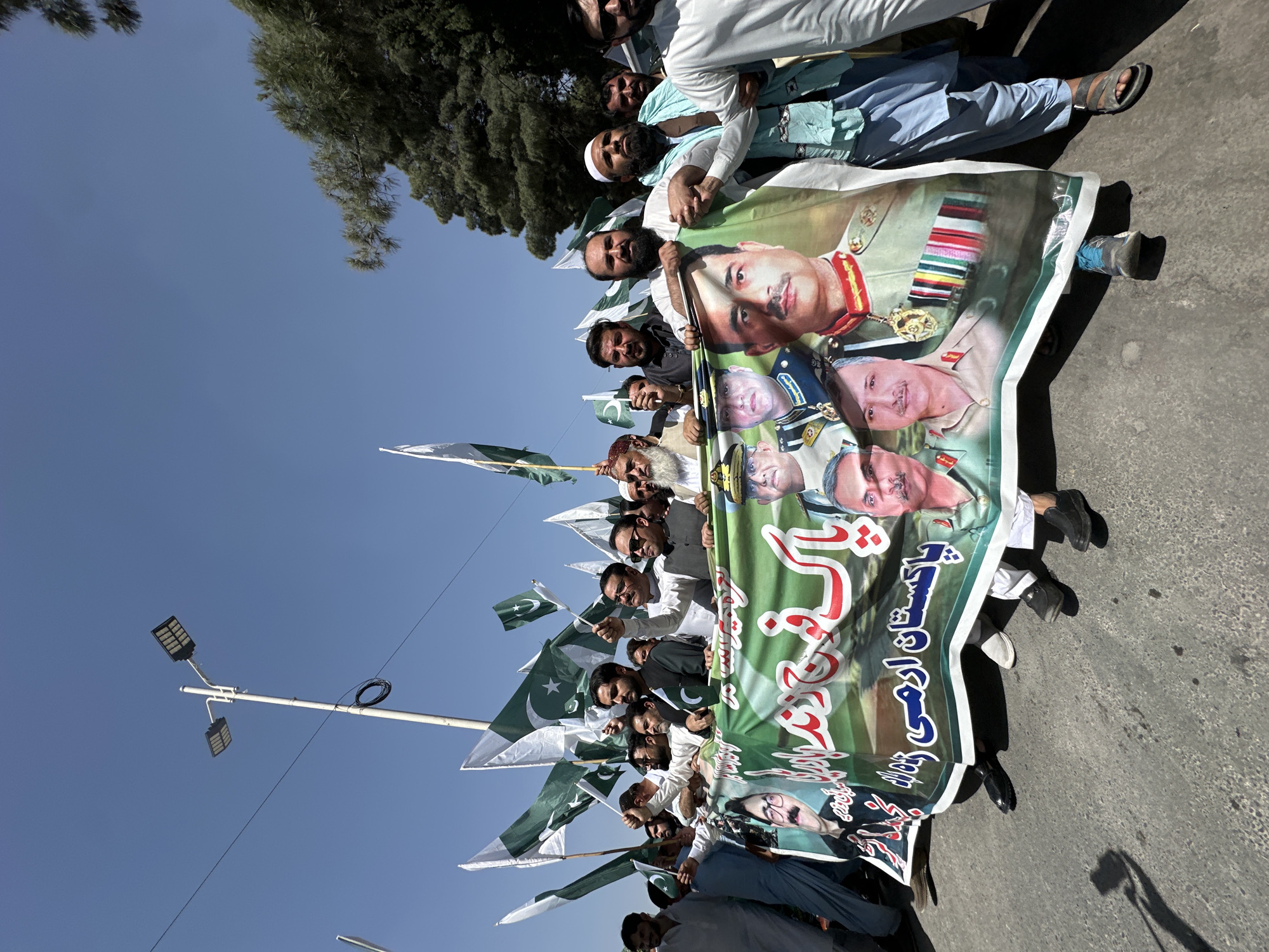 A medium shot depicts a group of men holding a large banner with the images of various military officers. The men are mostly wearing traditional Pakistani clothing, and some are holding Pakistani flags. The banner is green and white, with Urdu text. The background includes a blue sky, a tree, and a street lamp. The location is likely Quetta City Tehsil, Pakistan, as indicated by the flags and cultural context. The group appears to be participating in a rally or demonstration.