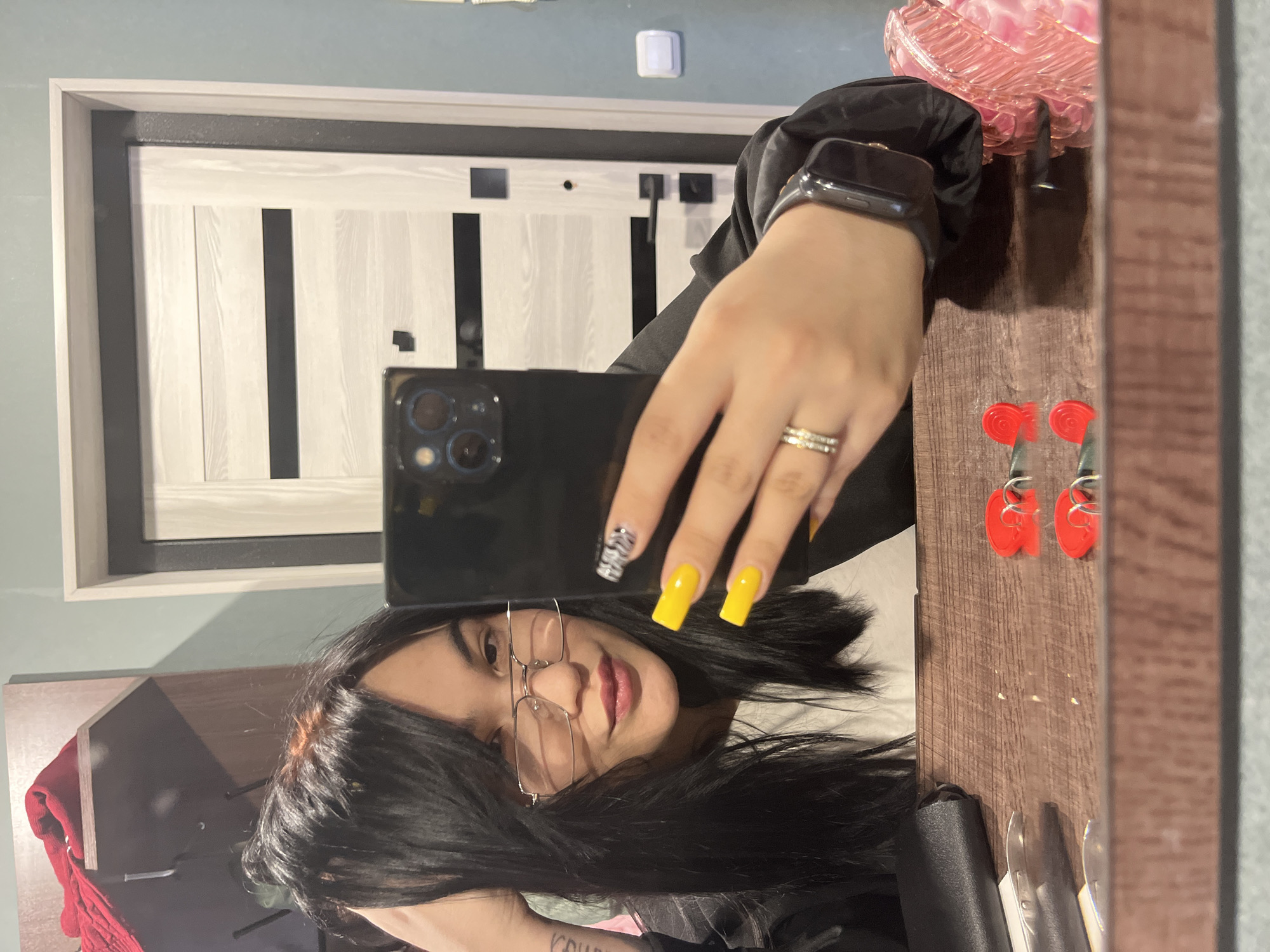 This media file contains a mirror selfie, originally rotated 90 degrees clockwise, featuring a young woman indoors.
**Subjects:**
* **Person:** A young woman with long, dark hair, wearing glasses and a black top. She is seen from the chest up, reflected in the mirror. She has light makeup, including pink lipstick, and a neutral to slightly smiling expression.
* **Objects:**
* **Smartphone:** A black smartphone, likely an iPhone, held by the woman, with a prominent triple-lens camera module visible.
* **Nails:** Her nails are long and square-shaped. Most are painted bright yellow, while one (on the hand holding the phone) has a black and white zebra print design.
* **Jewelry/Accessories:** She wears a multi-band ring on her ring finger and a dark-banded smartwatch on her wrist.
* **Furniture/Decor:** A modern door with a light wood or white finish and black vertical strips is in the background. A light blue/teal wall with a white light switch is visible above the door. A dark brown wooden surface, possibly a dresser or shelf, is to the right. On this surface are a pink, ridged, glass-like decorative object, two small red heart-shaped items with metal hooks (possibly earrings), and partially visible metal tools or cutlery. A red cloth or garment is reflected near the bottom left.
**Activity or event taking place:** The woman is taking a mirror selfie.
**Notable details:**
* **Setting:** The scene is an indoor residential environment, characterized by modern decor including the door and wall color. The wooden surface suggests a bedroom or hallway.
* **Lighting:** The lighting appears to be artificial and ample, illuminating the scene clearly.
* **Emotions:** The woman's expression is calm and composed.
**Visible text:** No legible text is visible in the image.