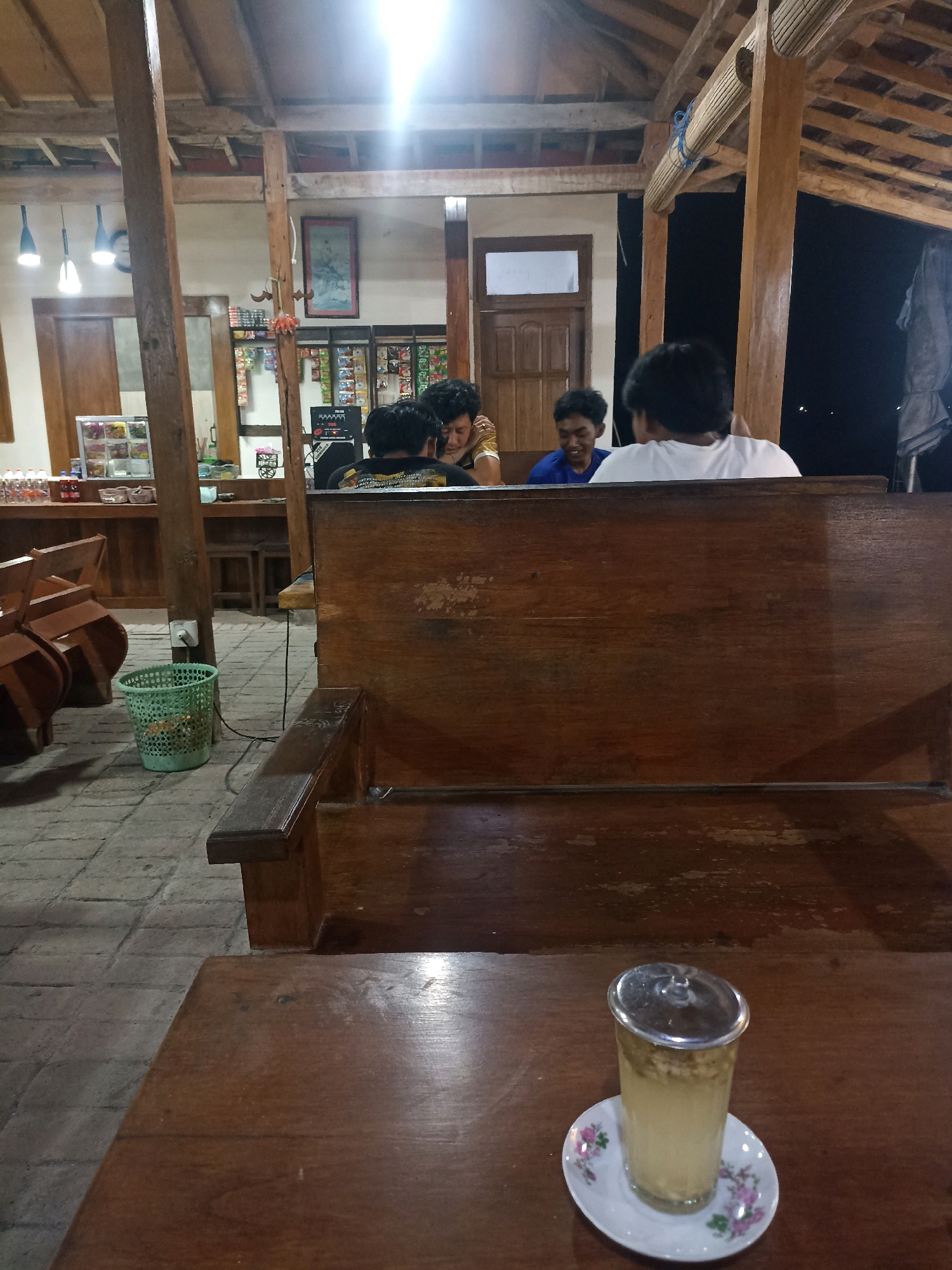 In the city of Bakalanpule, Indonesia, a group of individuals are seated at a wooden table in an open-air establishment, possibly a cafe or warung. The atmosphere is dimly lit, suggesting it is evening or nighttime. Several people are visible, including three men sitting together and another man in the foreground. One of the men is wearing a blue shirt, another a black t-shirt with yellow text, and a third is in a white shirt. The man in the foreground is facing away from the camera, only his back visible. A glass of beverage with a lid and a small plate with a floral pattern rest on the wooden table. The establishment features wooden beams and a thatched roof, typical of rural Indonesian architecture. In the background, shelves are stocked with various items, likely food and drinks. A green plastic trash can is situated near a wooden pillar, with a power cord plugged into a wall socket. The floor is paved with stone tiles. The overall scene depicts a casual gathering in a rustic, possibly outdoor, dining or seating area.