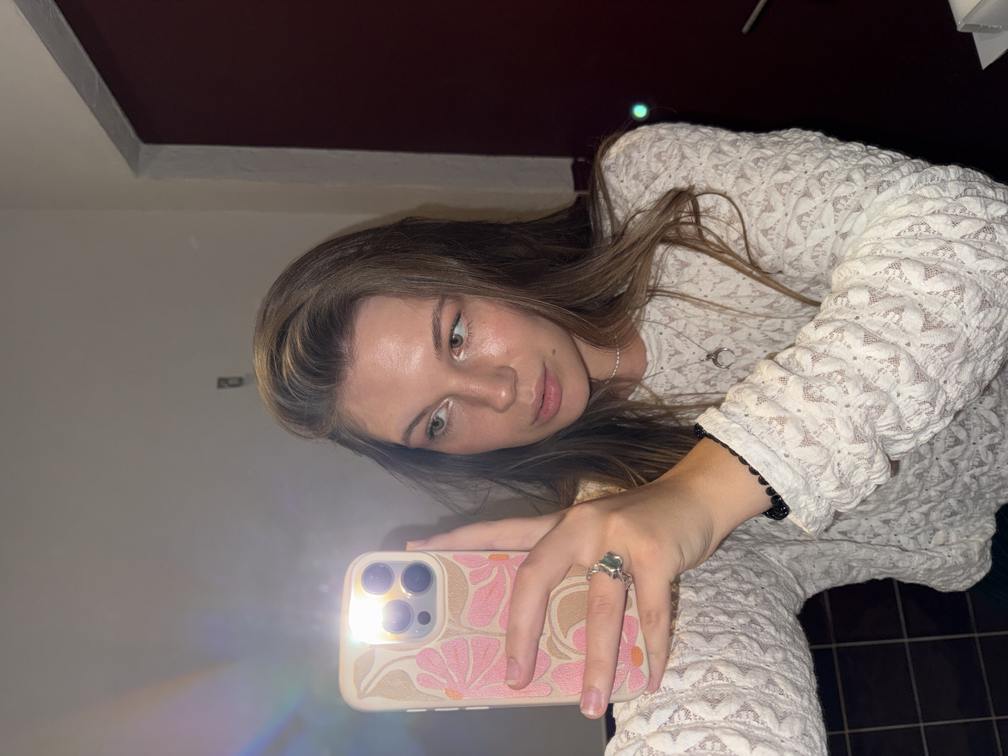 A young woman with long, light brown hair is taking a selfie in what appears to be a restroom. She is wearing a white lace top with a high neckline and a moon-shaped necklace. On her left hand, she wears a ring with a green stone. Her phone, with a floral-patterned case, is held up to take the picture, and the flash is illuminating her face and the surrounding area. Behind her, a dark red door is visible, and the walls are a light, neutral color. A rainbow-like light refraction is present in the upper left corner of the image. The floor appears to be tiled. There is no specific indication of the location being in Belgrade, Serbia, but the overall setting suggests an indoor, private space.