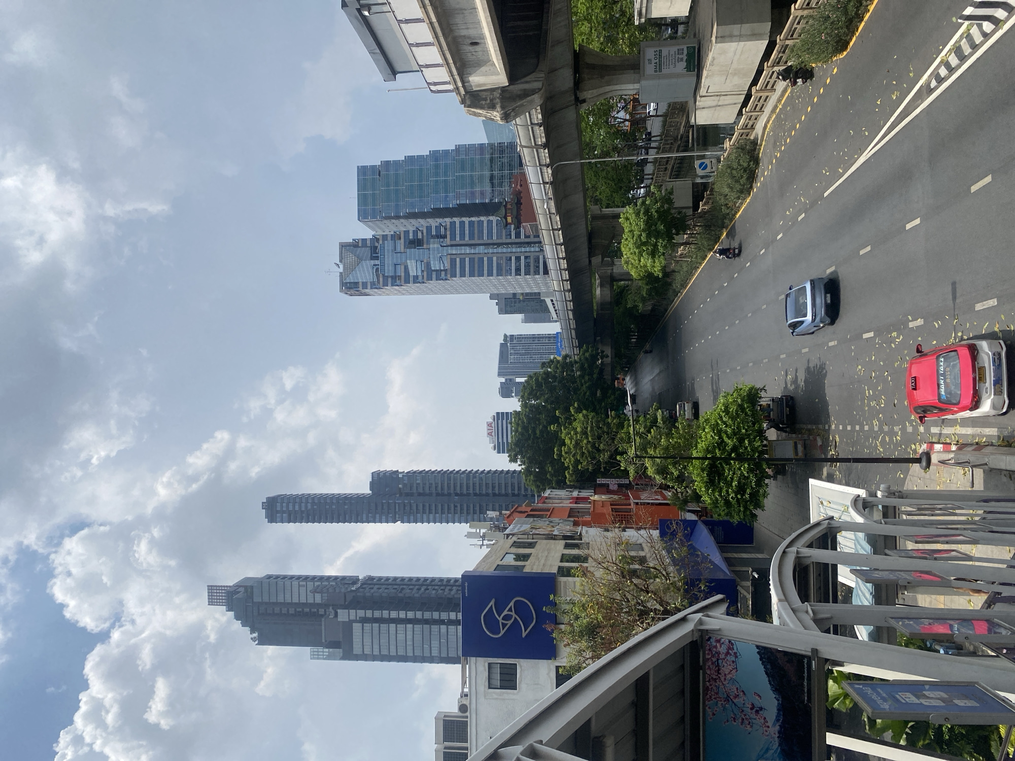 The image presents an elevated urban landscape in Bangkok, Thailand, captured from a pedestrian overpass or high vantage point. The sky is largely overcast with fluffy white and grey clouds, suggesting a partly cloudy day.
Dominating the scene are several modern skyscrapers with sleek glass facades, rising high above the city. Some exhibit contemporary architectural designs with recessed sections and varying window patterns. Below the towering structures, a mix of lower-rise commercial buildings with colorful accents and some greenery line the streets. On the left side, a concrete elevated railway track, characteristic of the BTS Skytrain system, runs horizontally across the midground, supported by sturdy pillars.
In the foreground and middle ground, an asphalt road with multiple lanes stretches through the scene. White solid and dashed lines mark the lanes, while yellow lines define shoulders and turns. Scattered yellow leaves are visible on the road surface, suggesting trees nearby. Vehicles populate the road, including a silver/blue car in the middle lane moving away from the viewer, and a distinctive red and white "SMART TAXI" on the right side. Another motorcycle is visible further down the road.
The immediate foreground on the right shows the metal railing and support structure of the viewpoint, below which advertising billboards are visible, one depicting a serene landscape with cherry blossoms. On a building below the elevated track, a large blue sign features a white, stylized "S" or swirling logo. Another white sign beneath the Skytrain track displays green QR codes and the text "BMA OSS" (likely Bangkok Metropolitan Administration One Stop Service), a clear indicator of the location. In the distance, on top of one of the skyscrapers, the letters "AIA" are faintly visible, likely part of a company logo. The overall atmosphere is that of a bustling, active city environment under a bright, though diffused, daylight.