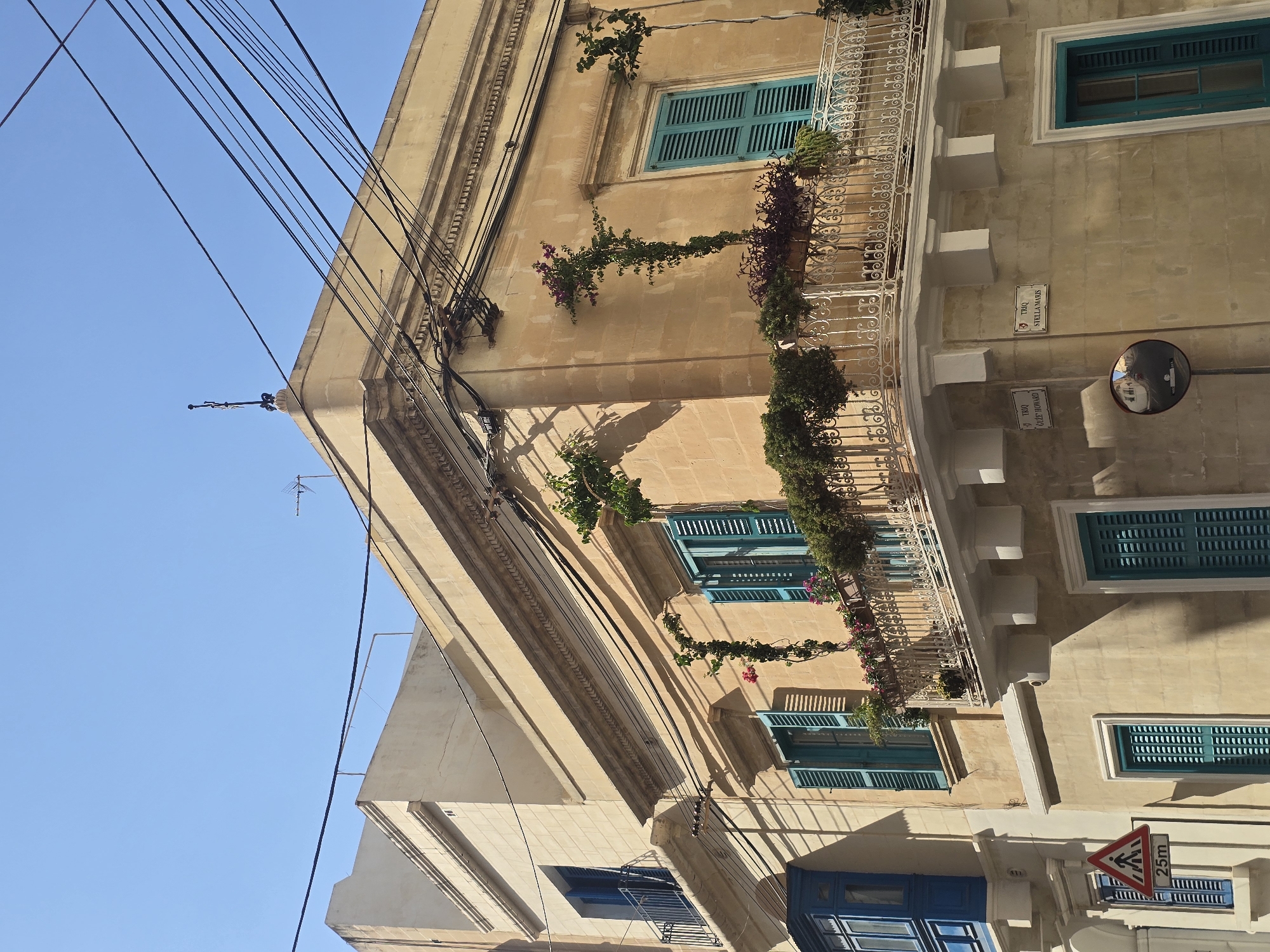 The image captures a close-up view of a building's facade in Sliema, Malta, under a clear blue sky. The building is constructed from light-colored stone, with architectural details like cornices and decorative mouldings. Multiple windows with teal-colored shutters are visible, some with wrought-iron balconies adorned with potted plants and trailing vines. The building features a prominent corner with a detailed white wrought-iron balcony on the upper floor, also decorated with greenery and flowers. Black electrical wires crisscross the sky above the building. In the lower left, a blue balcony with glass panels juts out. A street sign indicating "TRIQ GUZE HOWARD" and another reading "TRIQ STELLA MARIS" are affixed to the building's wall. A traffic mirror reflects a street scene. A pedestrian crossing sign with "25m" is also visible. The scene is bathed in bright sunlight, casting sharp shadows on the building. There are no people or animals present in the image.