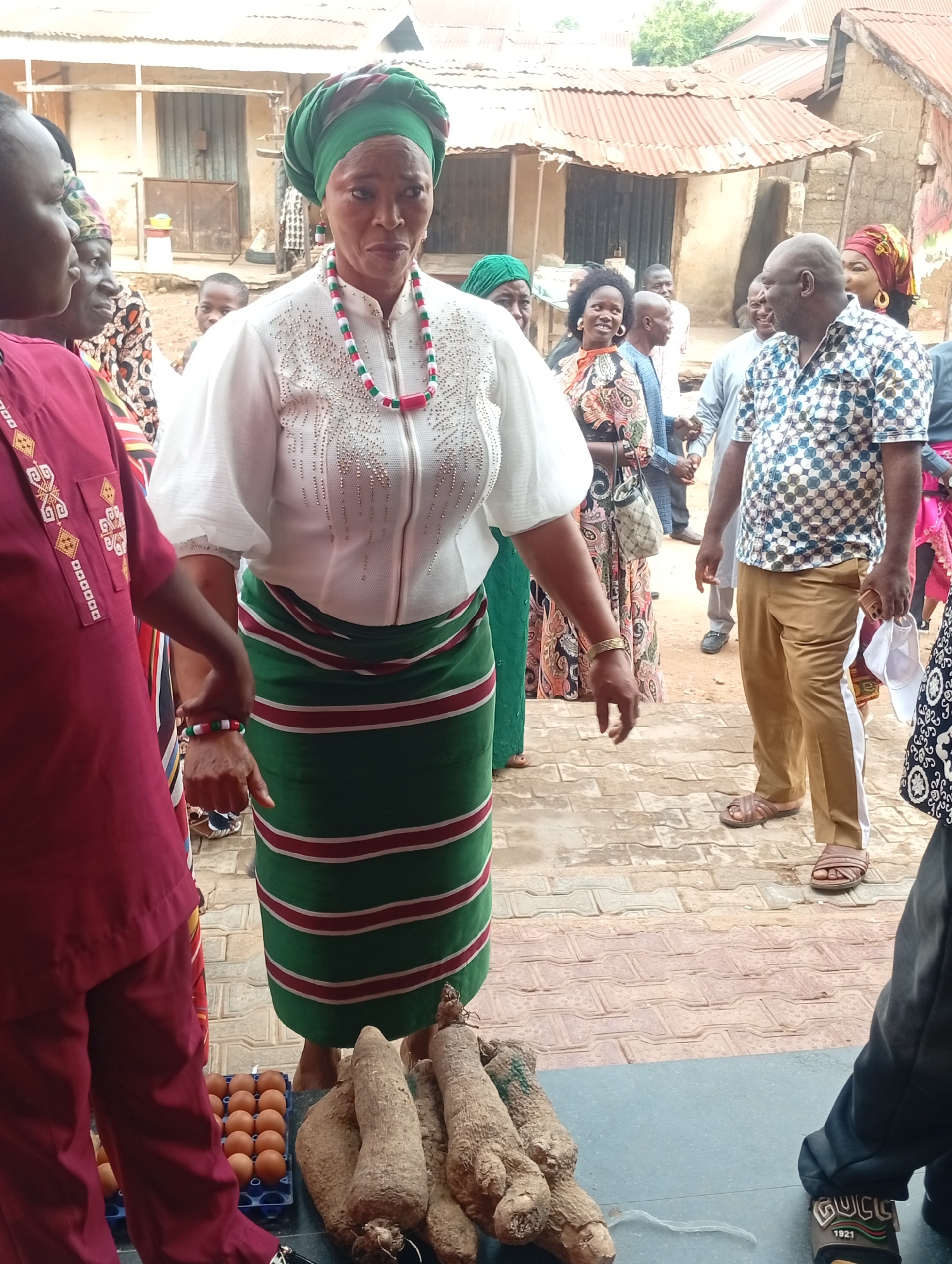 In Narayi, Nigeria, a woman in traditional Igbo attire—a green and white striped skirt, a white top with silver embellishments, and a green headwrap—stands next to a display of yams and a carton of eggs. She wears a necklace of red, green, and white beads. Surrounding her are people, some in traditional clothing, others in Western attire. A man in a patterned shirt and khaki pants stands nearby, and another man in a maroon outfit is partially visible on the left. The backdrop shows a corrugated metal roof and building. The scene appears to be outdoors, with patterned ground tiles visible.