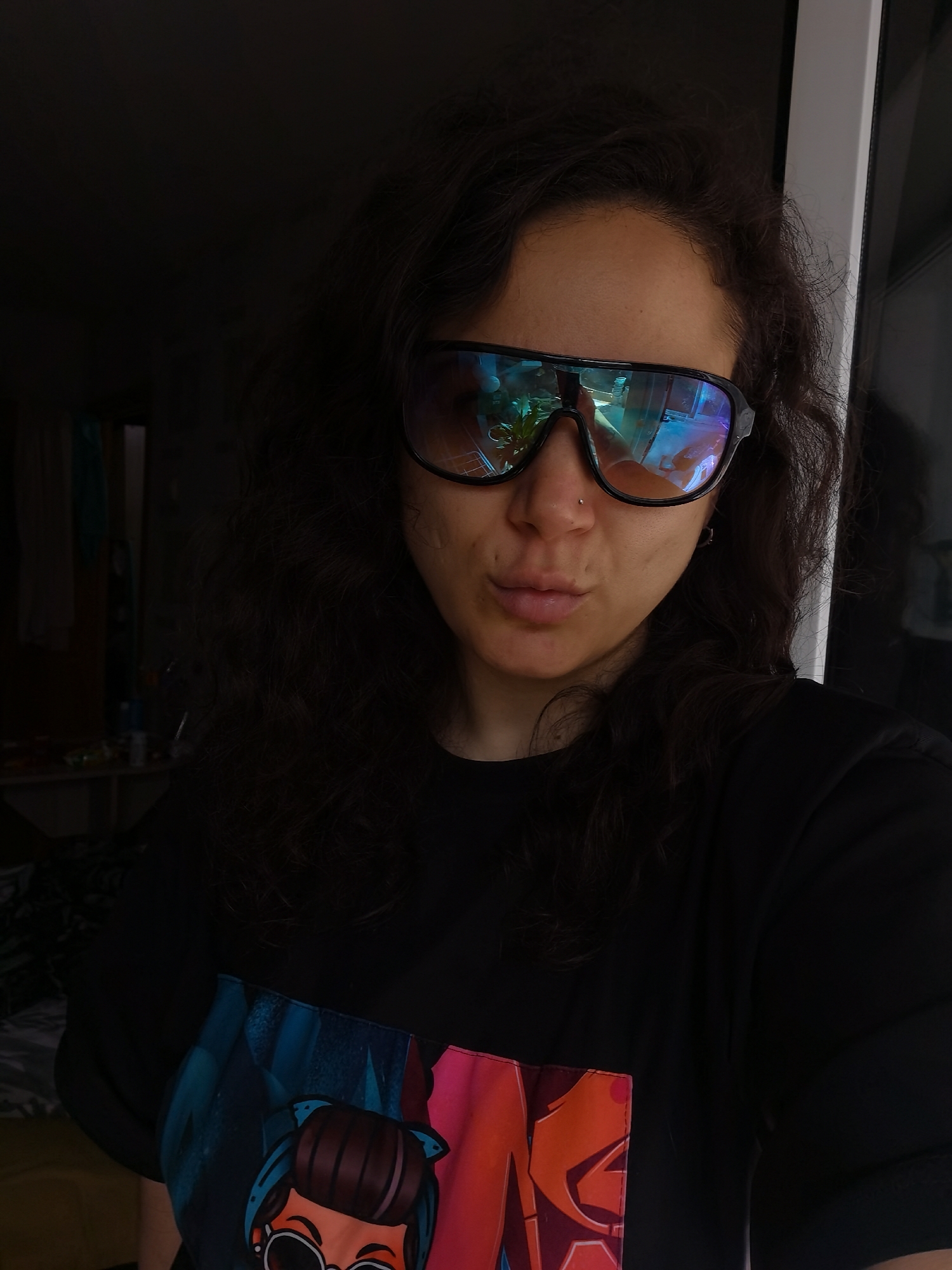 A young woman with dark, curly hair is taking a selfie in what appears to be a residential interior, possibly an apartment in Chaikovsky, Russia. She is wearing large, dark sunglasses with reflective lenses that show a distorted image of the room and a plant. She has a small nose ring and her lips are slightly pursed. She is wearing a black t-shirt with a colorful graphic print on the front, featuring a cartoon character with large glasses and blue pigtails. The background is dimly lit, with some shelving and possibly clothing visible on the left side of the frame. The overall lighting suggests an indoor setting, possibly near a window or doorway, with natural light contributing to the reflections on her sunglasses.