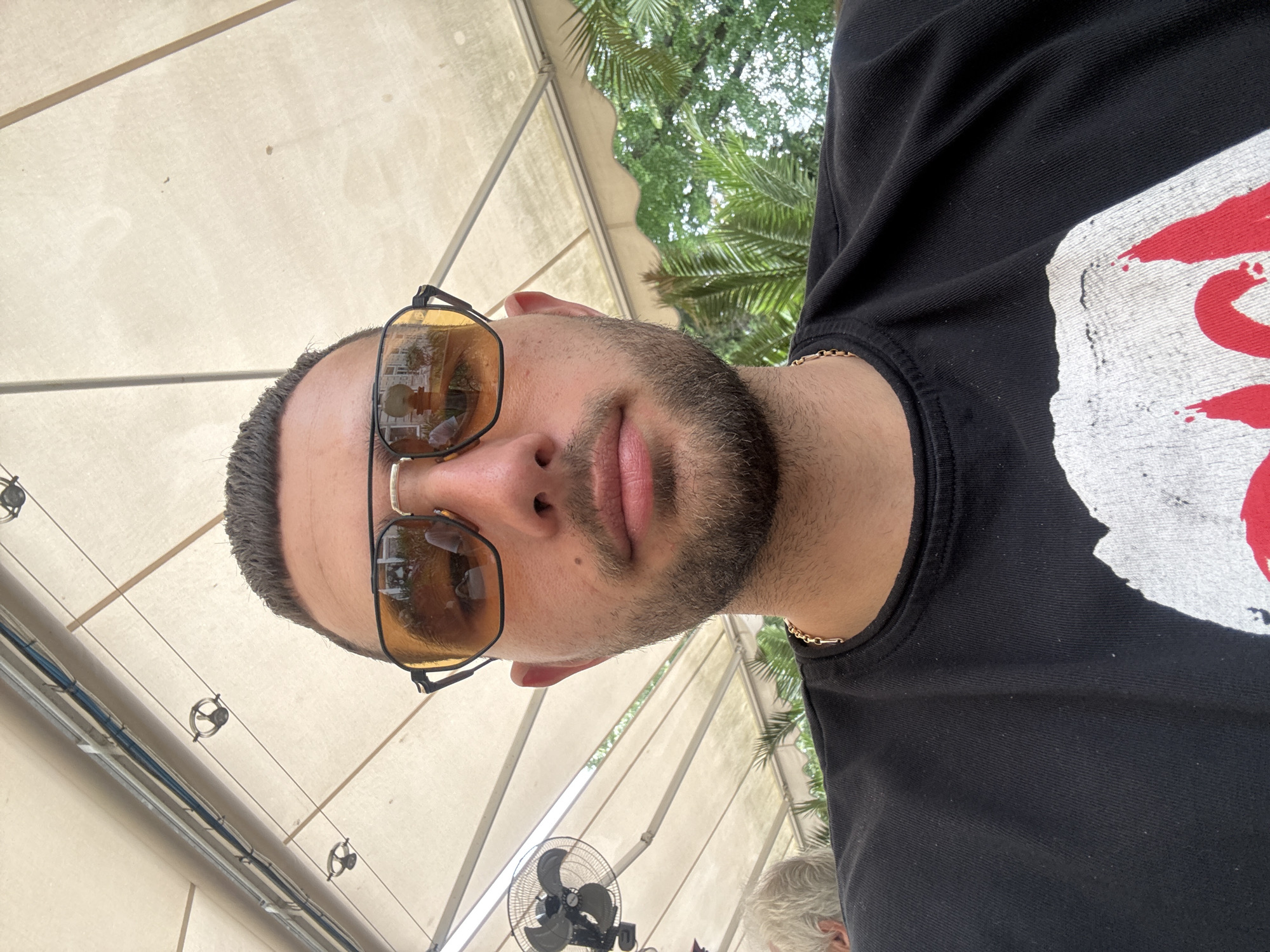 The image depicts a man with a beard and sunglasses, wearing a black t-shirt with a white and red graphic. He is located under a canvas awning, with palm trees visible in the background. A small fan is also visible. The location is Ruse, Bulgaria, as indicated by the reflection in the sunglasses, which shows a building with architecture typical of the city. The man is likely taking a selfie.
