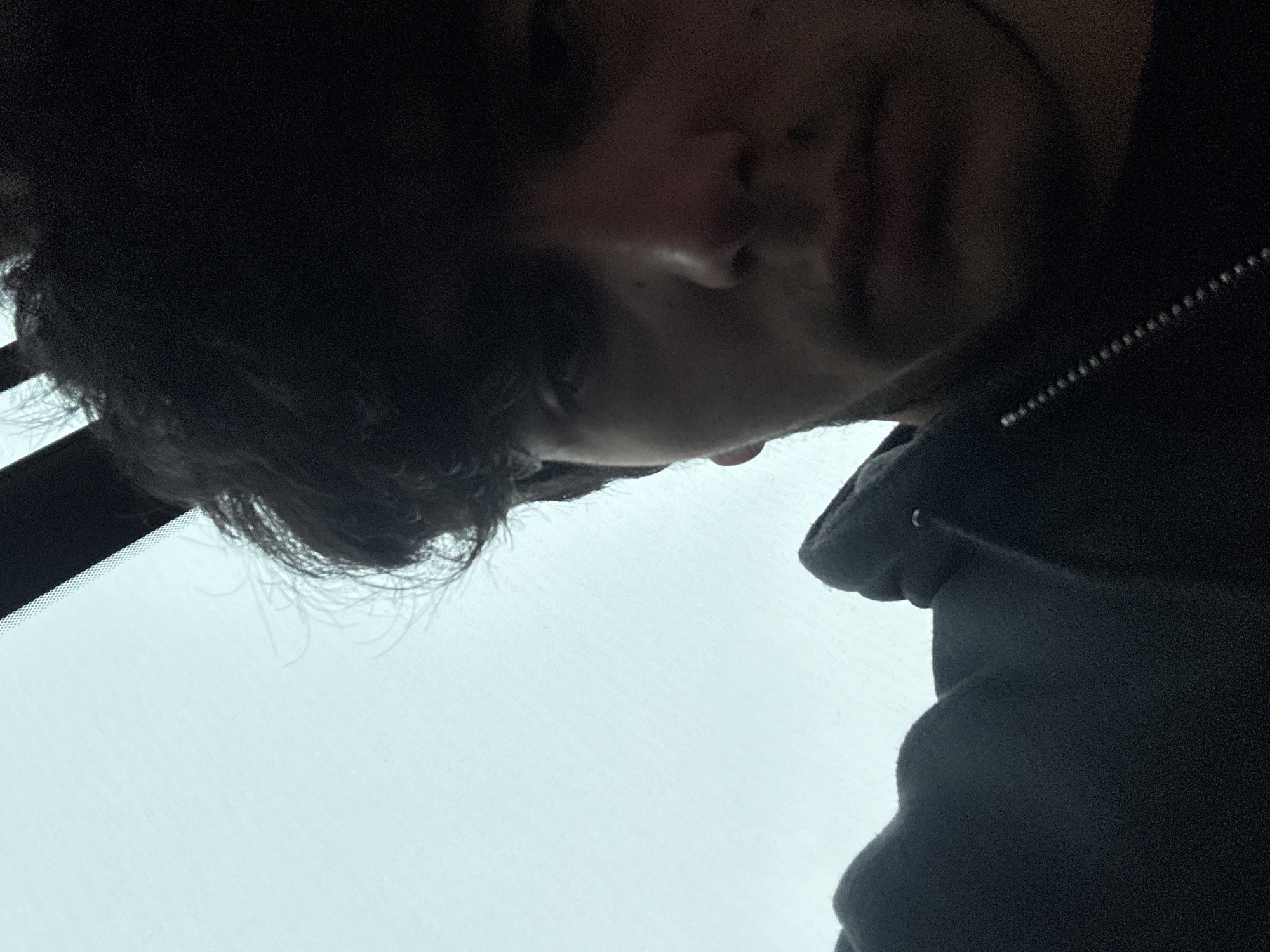 The image shows a person with dark hair and light skin. The person is wearing a dark-colored hooded jacket. The image appears to be taken from a low angle, possibly inside a car looking upwards toward the windshield. The background visible through the windshield is a light color, possibly the sky. The location is unknown.