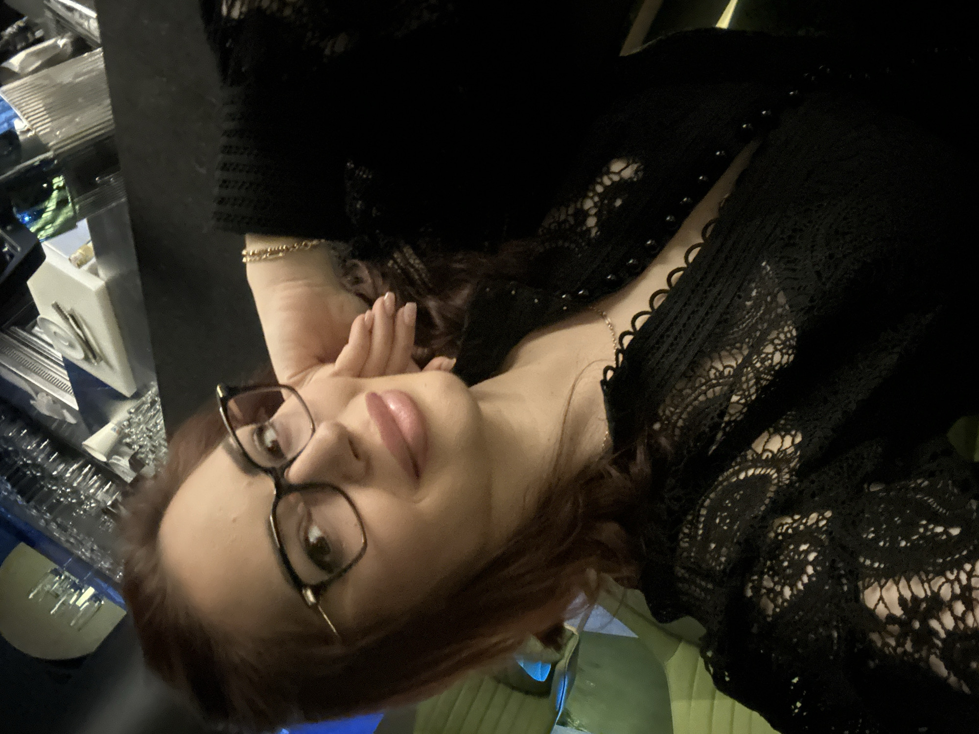 A woman with reddish-brown hair and dark-framed glasses is captured in a close-up, upward-angled shot within a dimly lit indoor environment, likely a bar or restaurant in Kazanlak, Bulgaria. She is wearing a black top or dress featuring intricate lace patterns, particularly visible on the sleeves and across the chest. A delicate chain necklace adorns her neck, and a thin, multi-strand gold-colored bracelet is worn on her right wrist. Her left hand, adorned with light-colored nail polish, is raised towards her face. Her lips are made up with light pink or nude lipstick, and she looks directly at the camera with a calm expression. The background is dark and blurry, showcasing reflective surfaces such as glass shelves or bottles, with subtle hints of blue and green light suggesting an evening or nighttime setting. The image is oriented sideways, presenting a tilted perspective. There is no visible text.