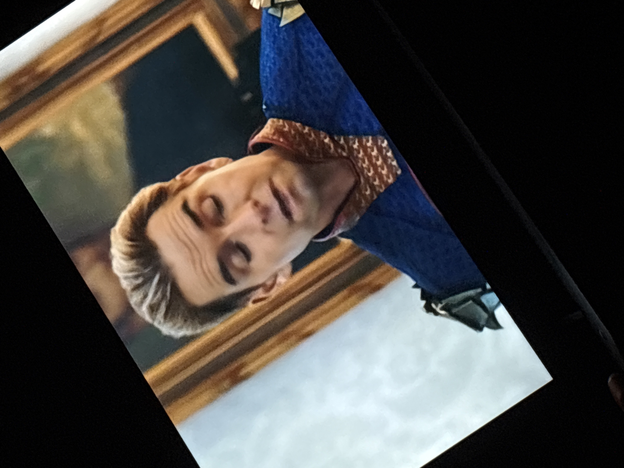 The image is a close-up, angled shot of a television screen displaying a scene from a show. The scene features a man with blonde hair, wearing a blue suit with gold and brown detailing around the collar and on the shoulders. He has a concerned or thoughtful expression. The background on the screen appears to be an interior setting with a large framed artwork. The television screen itself is set against a dark, out-of-focus background. No specific location in Dnipro, Ukraine, can be identified from this image.
