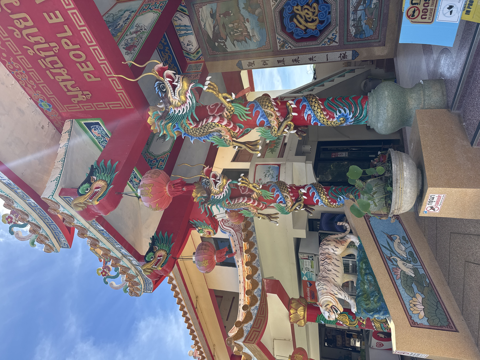 The image shows the exterior of a temple, likely in Baan Plai Laem, Thailand, given the presence of Thai script. The architecture is vibrant and ornate, featuring red and gold color schemes, intricate carvings, and traditional Chinese motifs like dragons. Two large, colorful dragon statues wrap around pillars, their scales detailed in blue, green, and gold. Red lanterns hang from the roof eaves, and decorative dragon heads protrude from the eaves as well. A statue of a tiger is positioned on a raised platform to the left, facing forward. The steps leading up to the temple are adorned with a mural depicting lotus flowers and cranes. Signs are visible, including one with Thai script and an image of bare feet with a prohibition symbol, indicating that shoes should be removed. The sky above is blue with scattered clouds. The overall scene conveys a sense of cultural richness and religious significance.