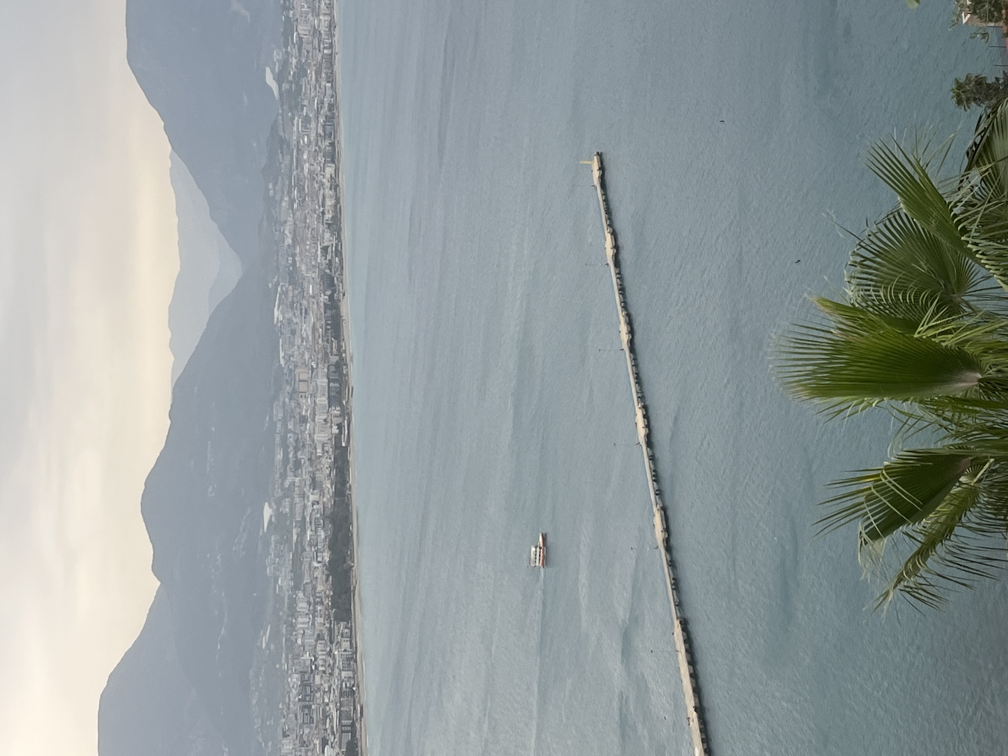 This aerial view captures a serene coastal scene in Alanya, Turkey. The image is dominated by the expansive, calm blue-grey sea on the right, stretching towards a long, light-colored pier or breakwater that extends far out from the coastline into the water. Along the left edge, layered, hazy blue-grey mountains rise, forming a dramatic backdrop to the densely built coastal city nestled at their base. Numerous buildings of the city can be seen sprawling along the shoreline.
On the sea, a small, white boat with distinct colored sections is visible, navigating the calm waters to the left of the long pier, suggesting ongoing maritime activity. In the upper right foreground, the vibrant green fronds of a palm tree partially frame the expansive vista.
The time of day appears to be daytime, characterized by a soft, diffused light and a slightly hazy sky over the mountains, possibly indicating an overcast day or early morning/late afternoon conditions. The weather is calm, with still water. No visible text is present in the image.