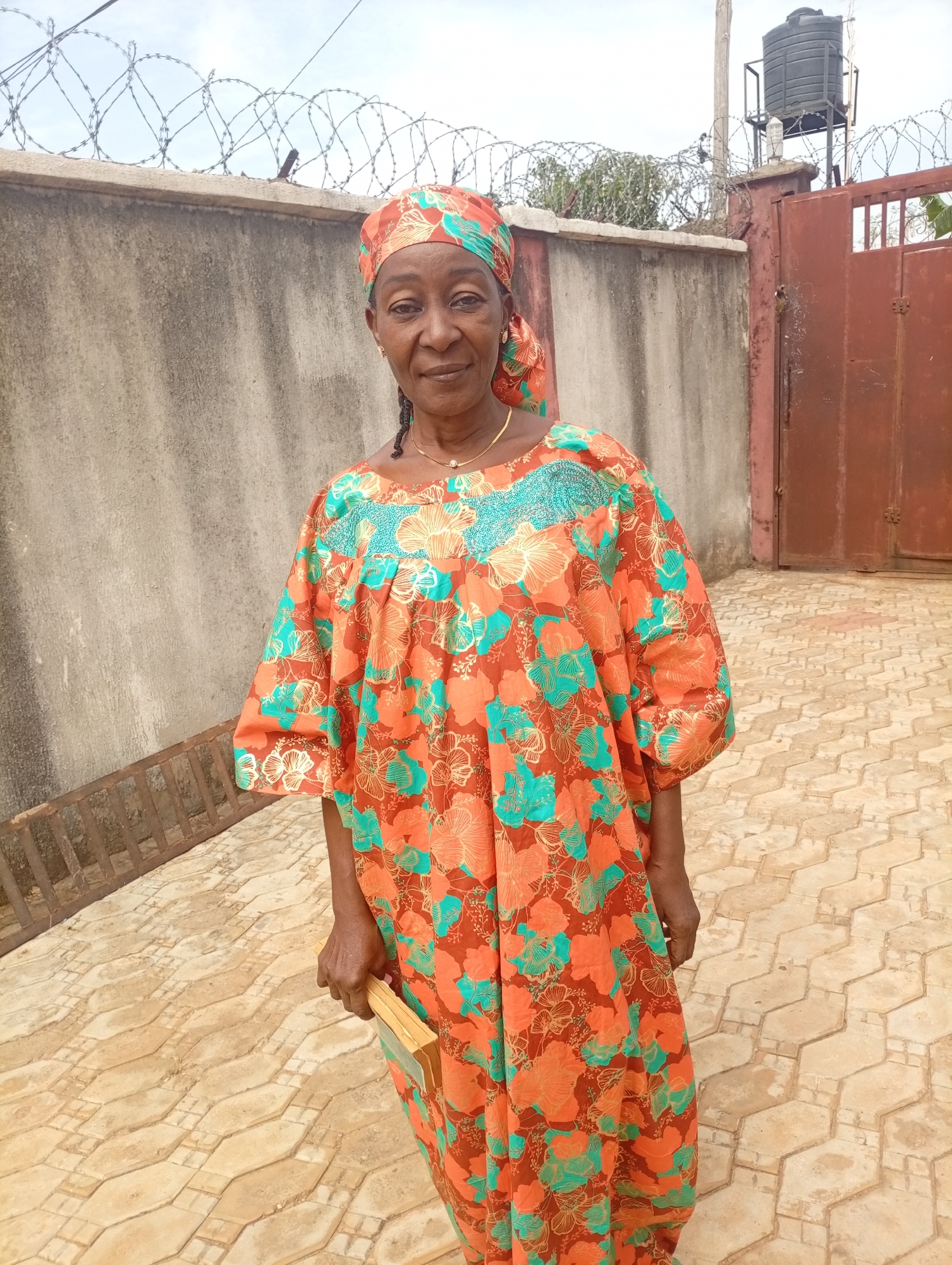 A woman stands outdoors in Rido, Nigeria. She wears a patterned dress and headscarf in shades of orange and teal. The background features a gray concrete wall topped with barbed wire, a brown gate, and a water tank on a post. The woman holds a small object in her left hand. The ground is covered in interlocking pavers.