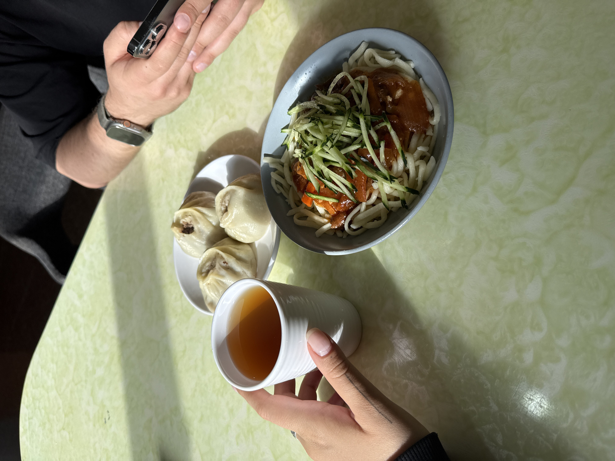 A top-down shot shows a shared meal on a green marble-patterned table. To the right of the frame, a grey bowl contains noodles topped with dark sauce and shredded cucumbers. To the left, a small white plate holds four dumplings. A white cup, containing a light brown liquid, is being held by a hand with a visible small tattoo. Another person is present; their hands are visible, holding a smartphone. The scene likely takes place in a restaurant or dining area, possibly located in Zabaykalsk, Russia, given the prompt's reference to that city.