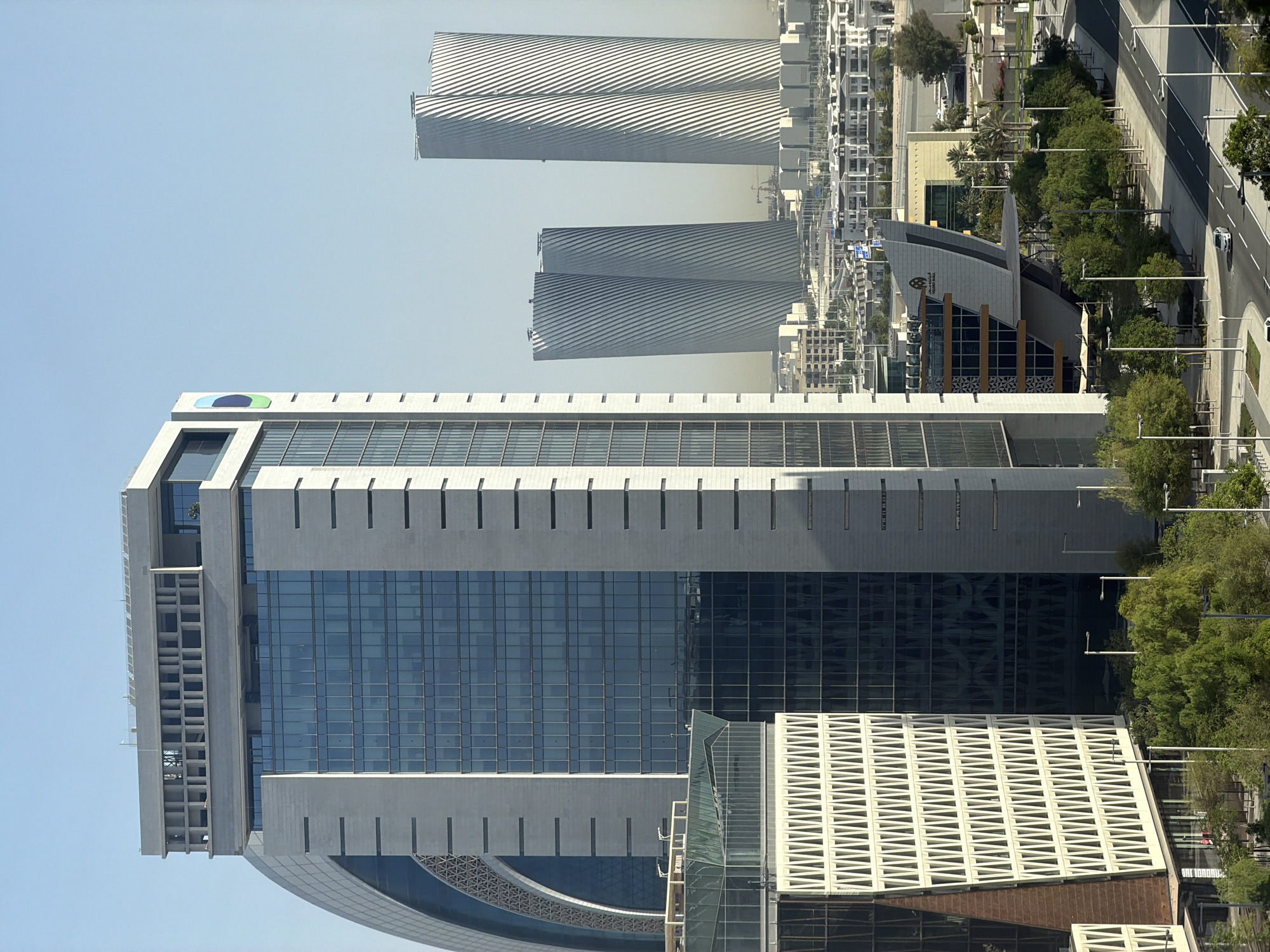 In an urban landscape bathed in clear, bright daylight, a cluster of modern skyscrapers dominates the scene. The tallest structure, a rectangular building with a facade of glass windows and light-colored stone panels, stands prominently in the foreground. Its upper section features a distinctive decorative element with a grid of rectangular openings. To its left, a curved building with a similar patterned facade is partially visible. In the midground, two strikingly tall, slender skyscrapers with a unique twisted, ribbed design rise into the sky. The surrounding urban environment consists of lower-rise buildings, trees, and roadways, with a few cars visible on the street. The sky above is a pale, uniform blue.