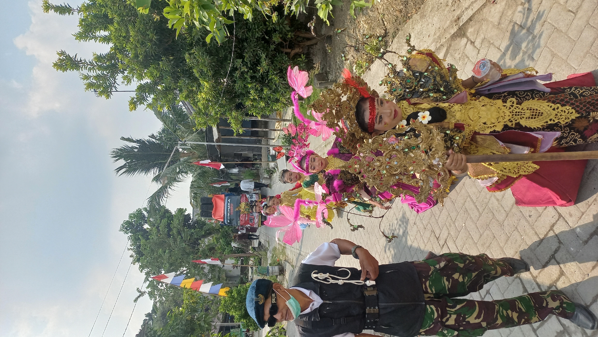In Kedungmegarih, Indonesia, a parade is underway. In the foreground, a man in military-style camouflage pants and a black vest, wearing sunglasses and a light blue mask, walks to the left of the frame. He is followed by a group of children in ornate costumes. The children are dressed in vibrant, gold and pink traditional outfits, with elaborate headdresses and wing-like accessories. One child in the front is holding a tall, gold decorative staff. Behind the children, a truck with speakers is visible, suggesting a festive or celebratory event. Indonesian flags are displayed along the street, indicating a national holiday or commemoration. The scene takes place on a paved street, with trees and buildings lining the sides, under a bright, partly cloudy sky.