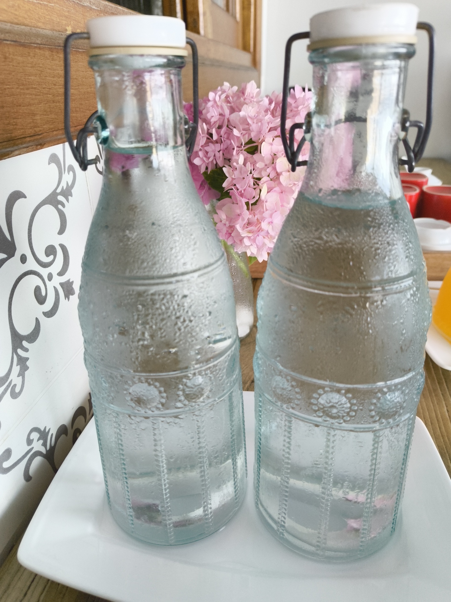 This is a close-up, eye-level, indoor shot of two glass bottles filled with clear liquid. The bottles are cylindrical with textured patterns of raised dots and vertical lines. Both bottles have white stoppers with black wire closures and beige rubber seals. The bottle on the left is slightly closer to the camera and positioned in the foreground. The bottle on the right is slightly behind and to the right of the first. The bottles are sitting on a white rectangular plate, which is on a wooden surface. In the background, a bouquet of pink flowers is visible to the left of the bottles, and beyond that, a wooden surface, possibly furniture, and a portion of a tiled wall with a gray ornate pattern. The lighting is bright, creating reflections and condensation on the glass bottles. There are no people or animals visible in the image. The location is not specified as Phu Thuy Ward, Vietnam.