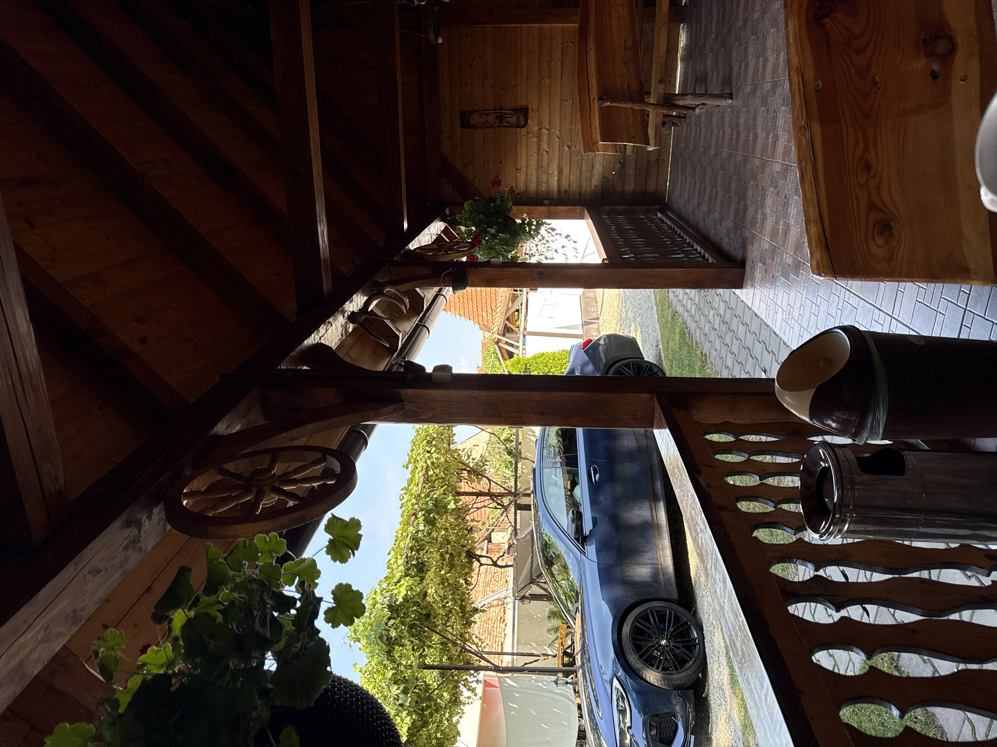 This image depicts an outdoor covered patio with wooden construction and furnishings. A blue car is parked in front of the patio, and a wooded area with trees and a brick building can be seen in the background. The patio has a wooden railing, a tiled floor, and wooden benches. Decorative elements like a wooden wheel and hanging plants are visible. The scene is likely located in Cârțișoara, Romania, given the architectural style and surrounding environment.