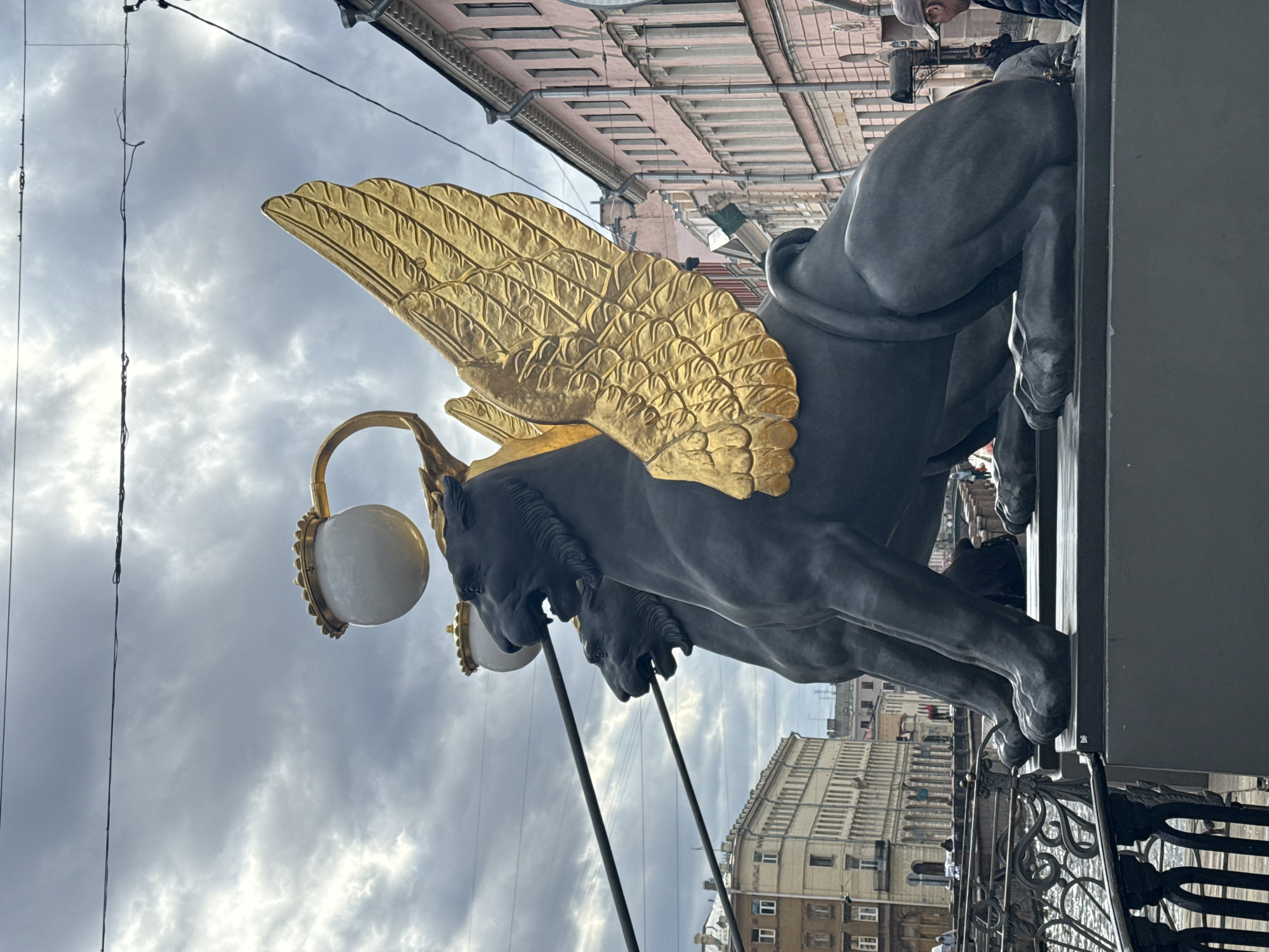 The image depicts a winged lion statue gracing the Bank Bridge in Saint Petersburg, Russia. The sculpture, rendered in a dark, matte material, features prominent golden wings. A lamppost with spherical globes stands adjacent to the statue. The background includes other architectural structures under a sky filled with gray, overcast clouds. The scene is captured from a low angle, emphasizing the statue and its details.